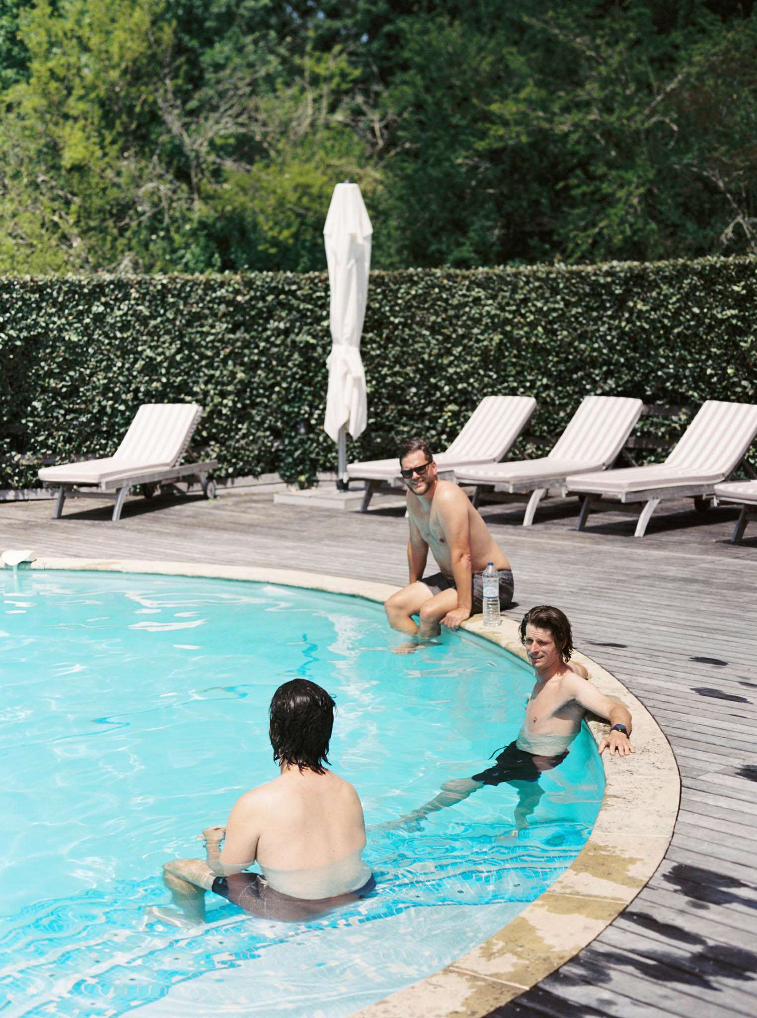 Three male wedding guests relax at an outdoor pool during what appears to be a pre- or post-wedding leisure period. Two men are in the water while a third sits on the pool edge with feet in the pool, wearing sunglasses and smiling toward the camera. The pool has a curved shape with a stone coping edge and a wooden deck surround lined with cream and white striped sun loungers and a closed white parasol. A neatly trimmed hedgerow borders the pool area. The shot is taken from a slightly elevated angle, giving a wide view of the pool and its surroundings.