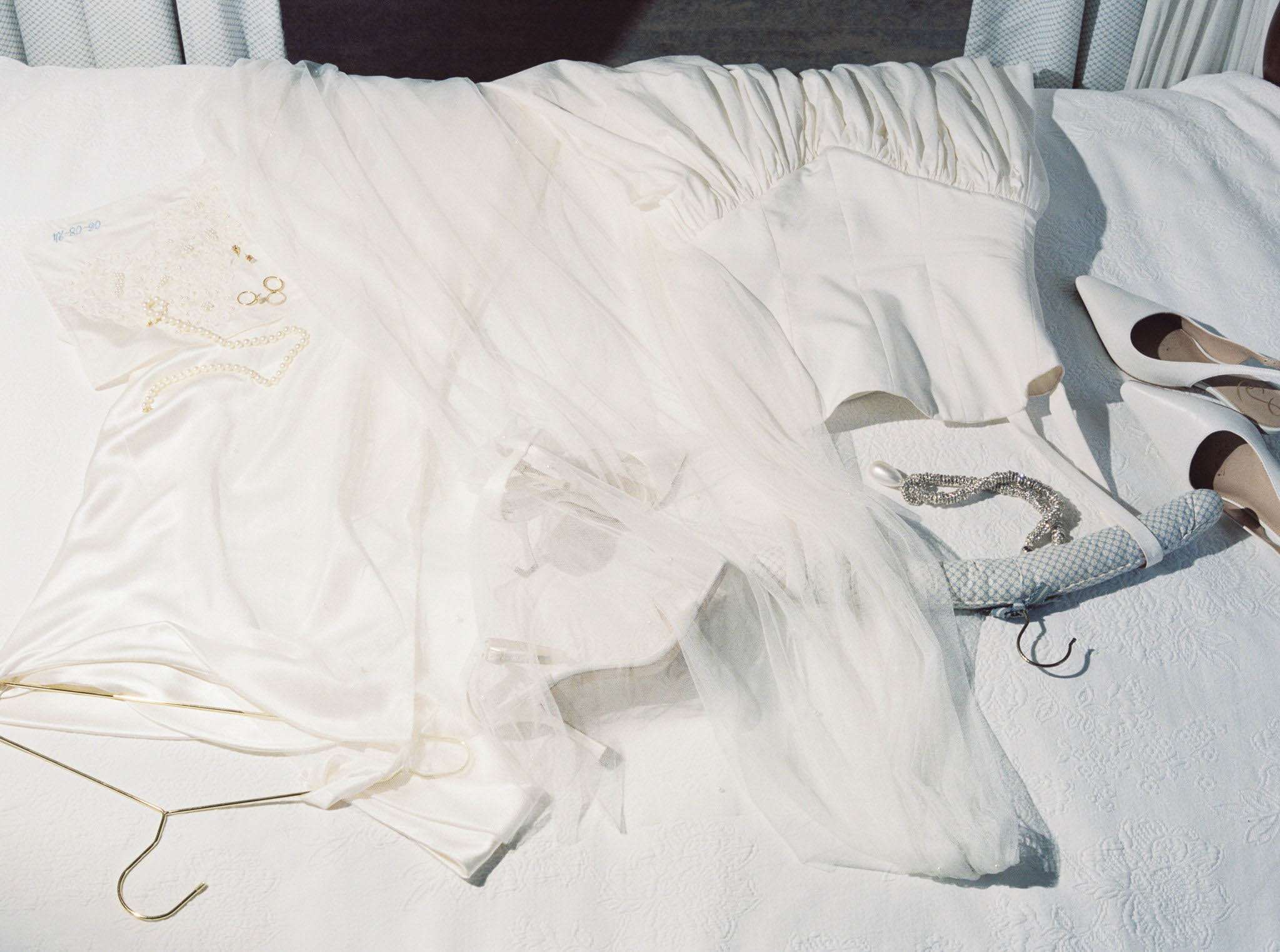 A bridal flat-lay detail shot of wedding accessories arranged on a white textured bedspread in a hotel or getting-ready room. The ivory wedding dress skirt with a tulle overlay and gathered waistband is spread across the bed, accompanied by a gold wire dress hanger at the bottom left. To the upper left, a small lace fabric swatch holds gold hoop earrings and a delicate pearl necklace. On the right side, two pairs of shoes are visible: white satin pointed-toe heels with dark cap toes, and pale blue quilted ballet flats with a crystal-embellished ankle strap. Strong natural light casts sharp shadows across the scene, giving the image a bright, high-contrast daytime look.