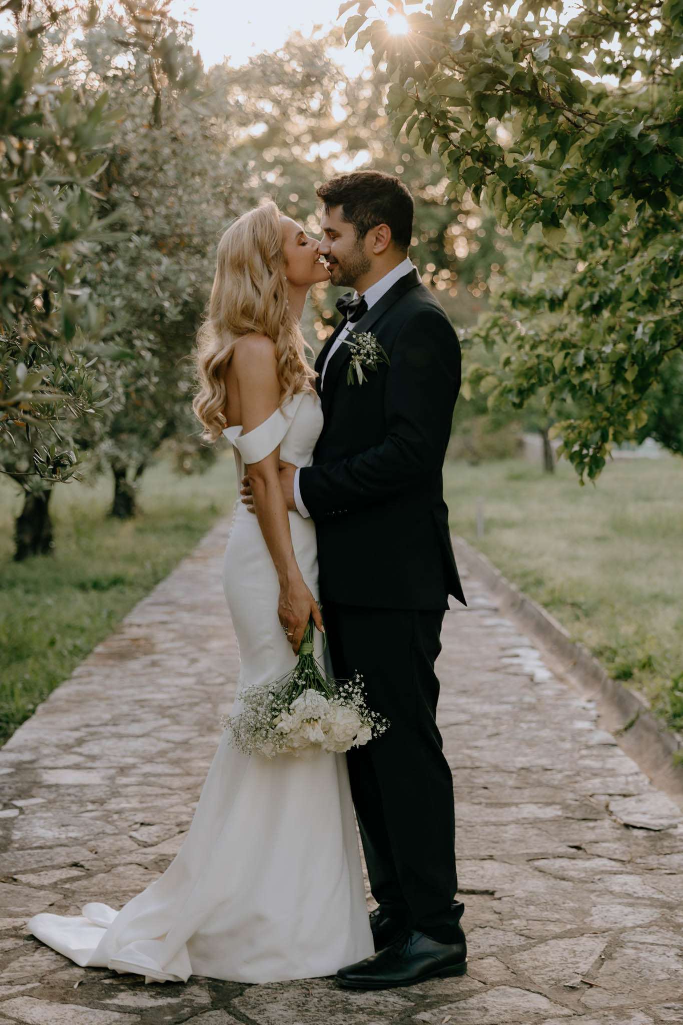 Bride with peony bouquet and groom in tuxedo lean in to kiss on olive-tree-lined path at golden hour