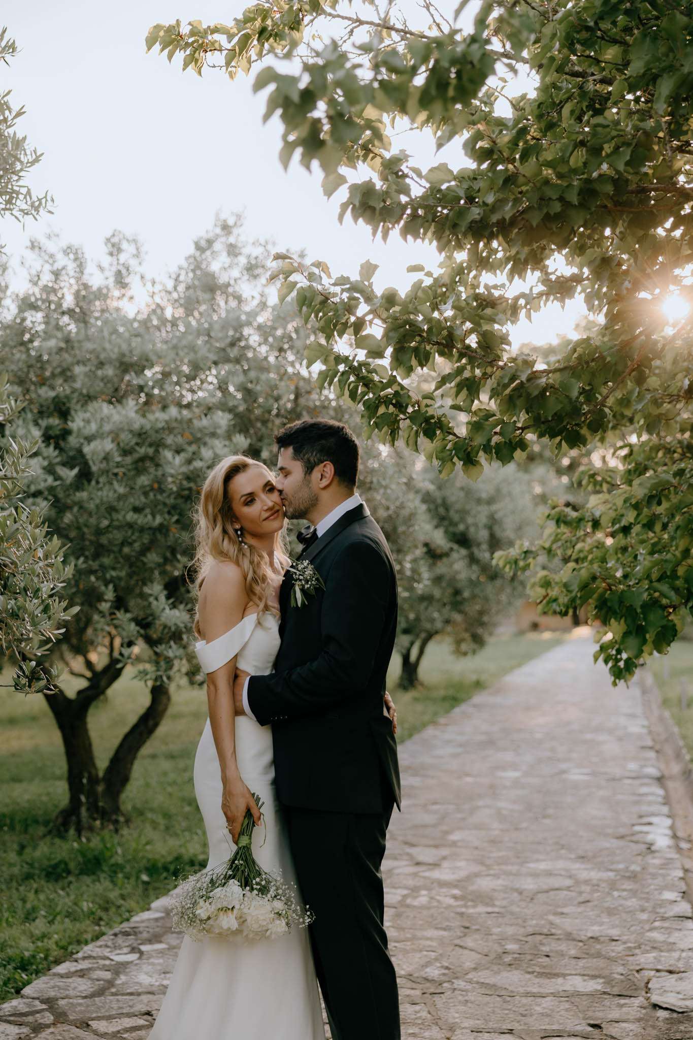 Groom kissing brides cheek on olive tree path at golden hour bride holding white peony and babys breath bouquet