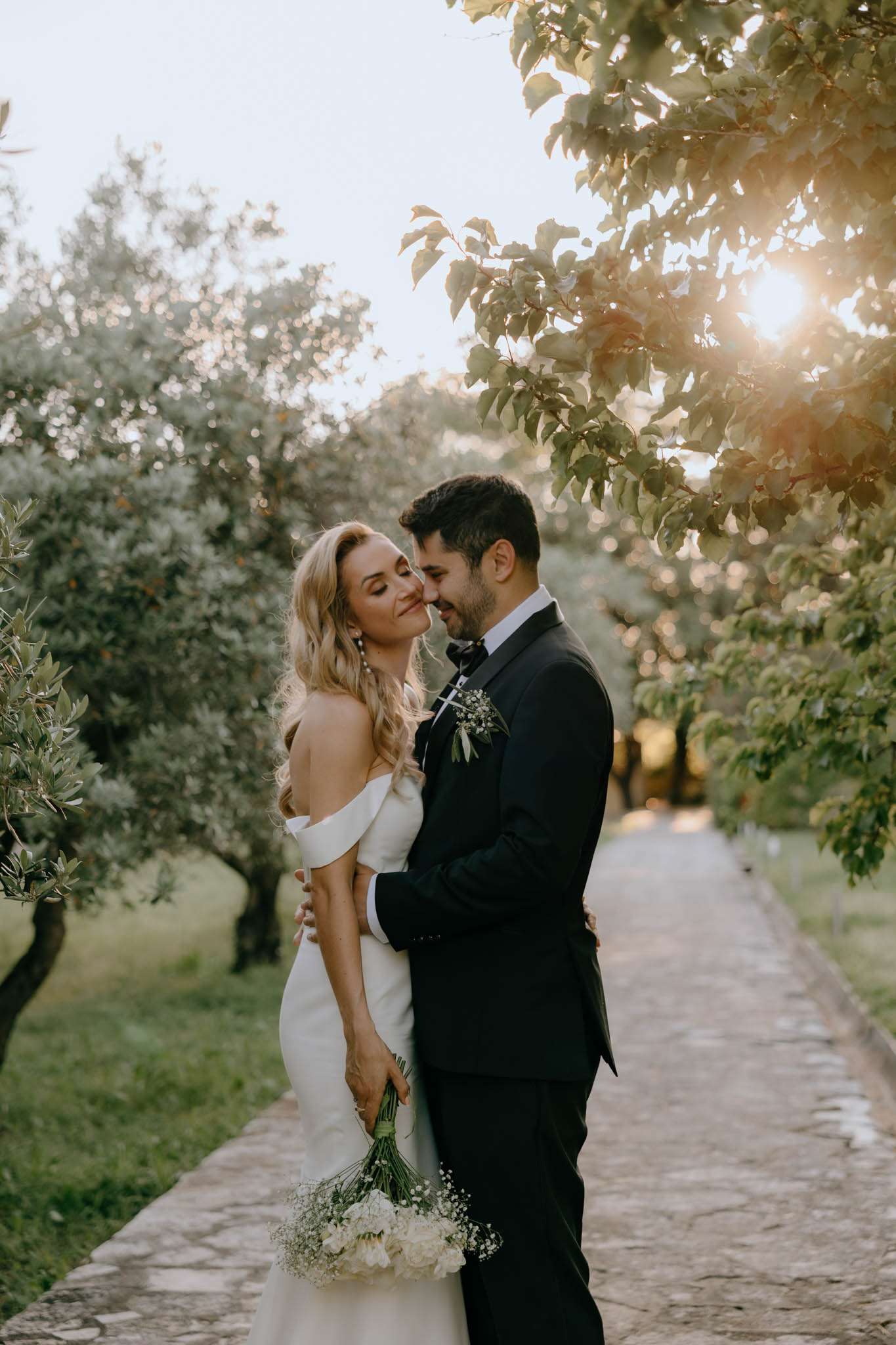 Close-up couple foreheads touching on olive tree path at golden hour bride with white rose bouquet groom in tuxedo