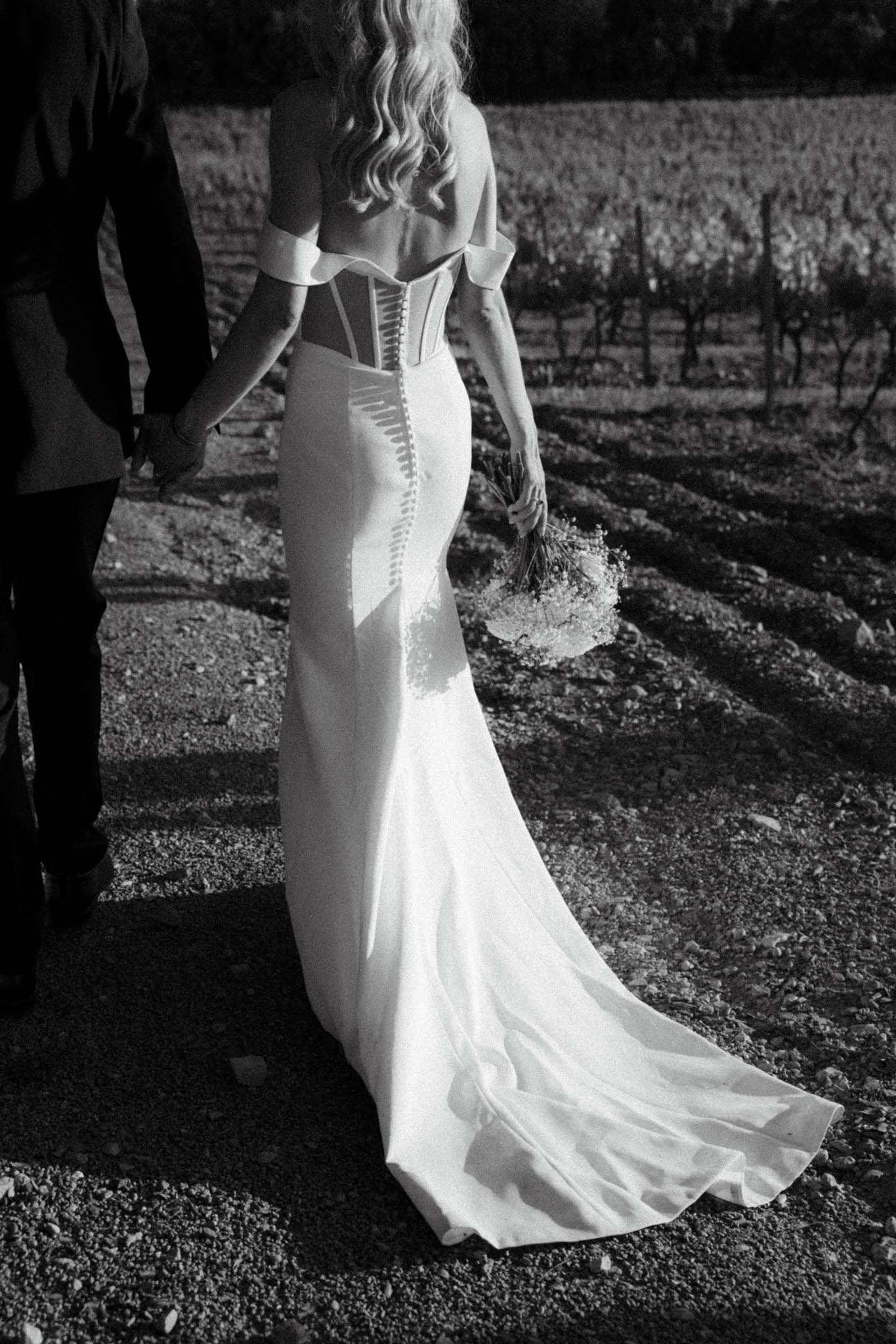 This black-and-white portrait captures the bride and groom from behind during what appears to be a couples portrait session at a vineyard. The bride wears a fitted off-the-shoulder mermaid gown with a structured corset back featuring detailed boning and a row of buttons running down the spine, along with a flowing train that trails on the gravel path. Her wavy blonde hair falls loosely down her back, and she holds a small rounded bouquet at her side. The groom, partially visible on the left, is dressed in a dark suit. Strong directional sunlight creates sharp shadows across the ground and the bride's dress, producing high contrast tones throughout the image. The vineyard rows are clearly visible in the background, establishing an outdoor, rural French setting.