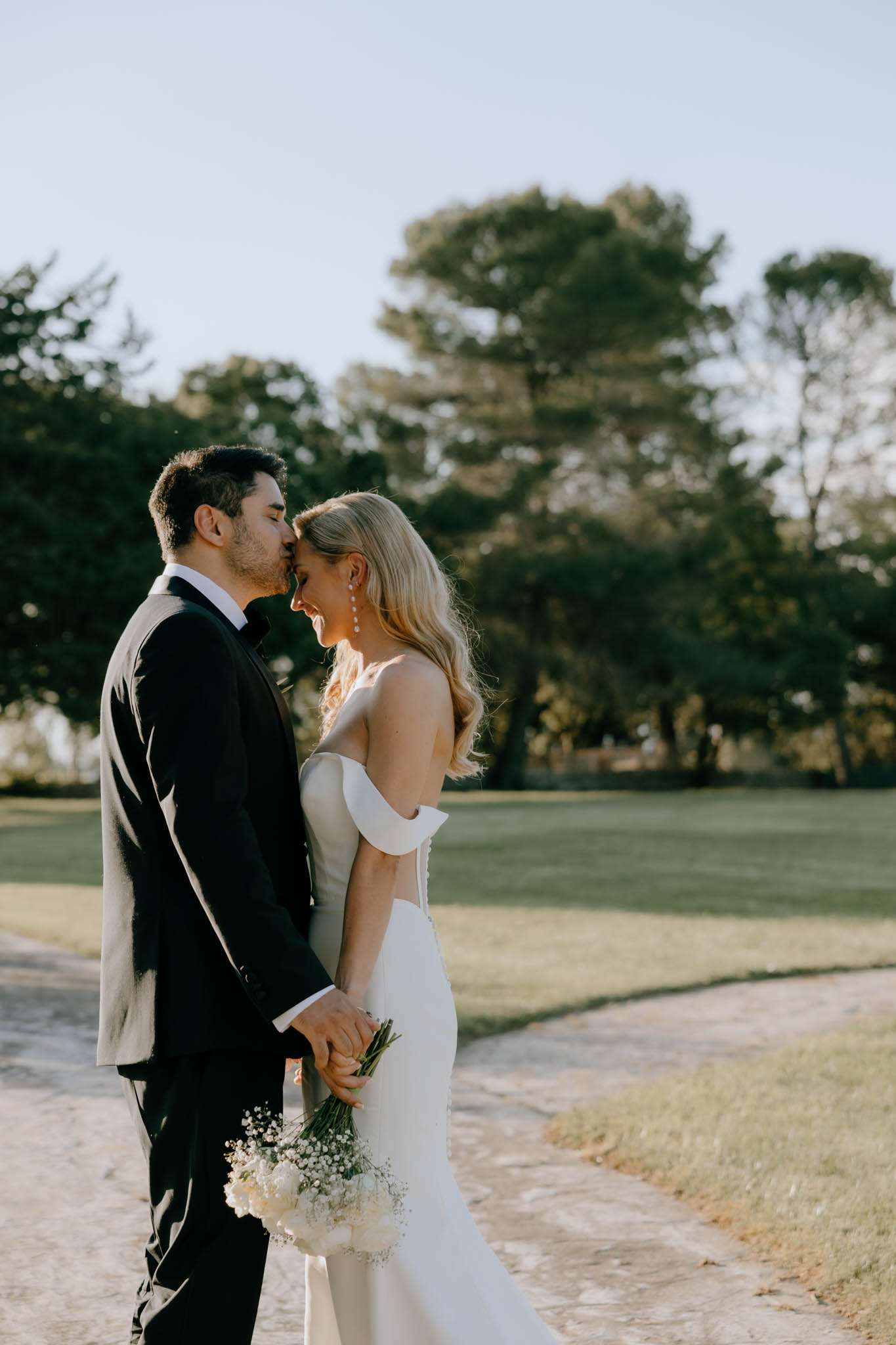 A couple portrait taken outdoors on a stone pathway, with the groom kissing the bride on the cheek as she laughs. The groom wears a black tuxedo with a bow tie, and the bride wears an ivory off-the-shoulder fitted gown with a low back and a small train. She holds a compact bouquet of white baby's breath and carries drop earrings. The overall styling is clean and modern. The shot is a medium portrait framed from roughly the knees up, with a soft-focus background of open lawn and mature trees in warm natural light.