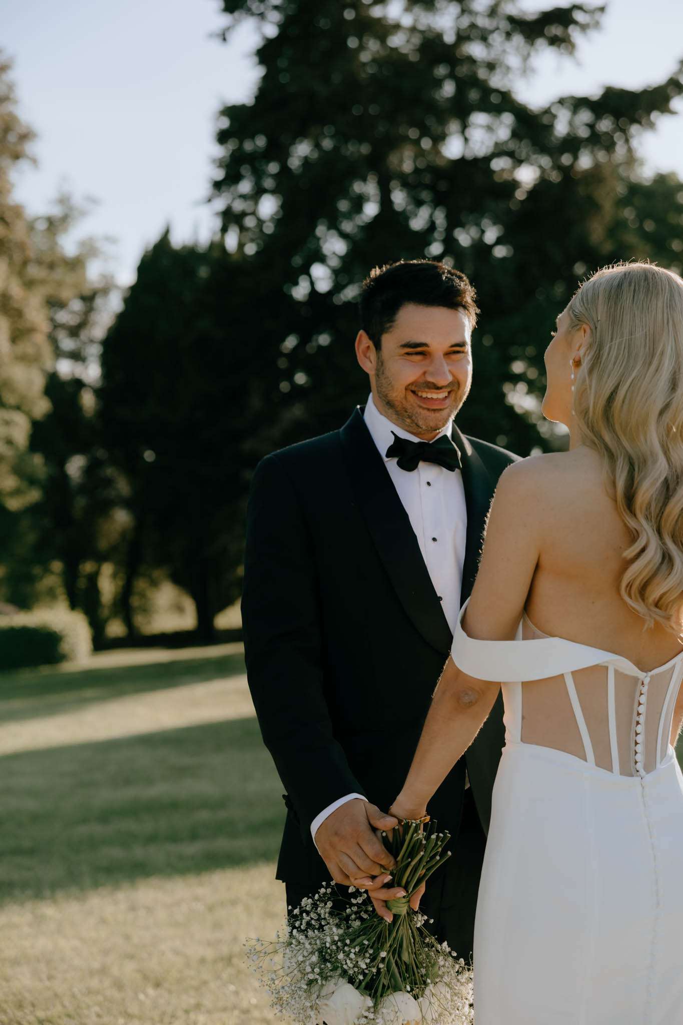 Groom in black tuxedo smiling at bride seen from behind in corset-back gown holding white gypsophila bouquet
