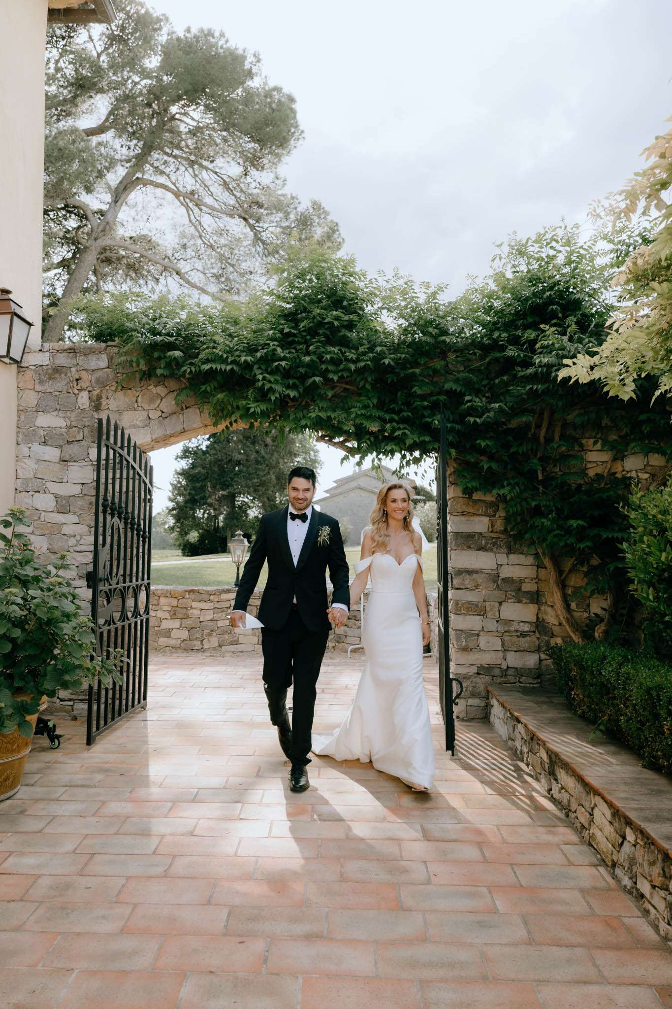 A couple walks hand-in-hand through an open wrought-iron gate set into a stone wall draped with climbing green foliage, framing them as they move toward the camera. The groom wears a black tuxedo with a black bow tie and a small boutonniere on his lapel, while the bride wears a fitted off-the-shoulder ivory satin gown with a sweetheart neckline and a trailing skirt. The setting is an outdoor terrace paved with warm terracotta tiles at a Provençal-style property featuring dressed stone architecture, a wall-mounted lantern, and manicured boxwood shrubs. The shot is a full-length portrait taken at eye level, with a stone building and open grounds visible through the gate in the background.