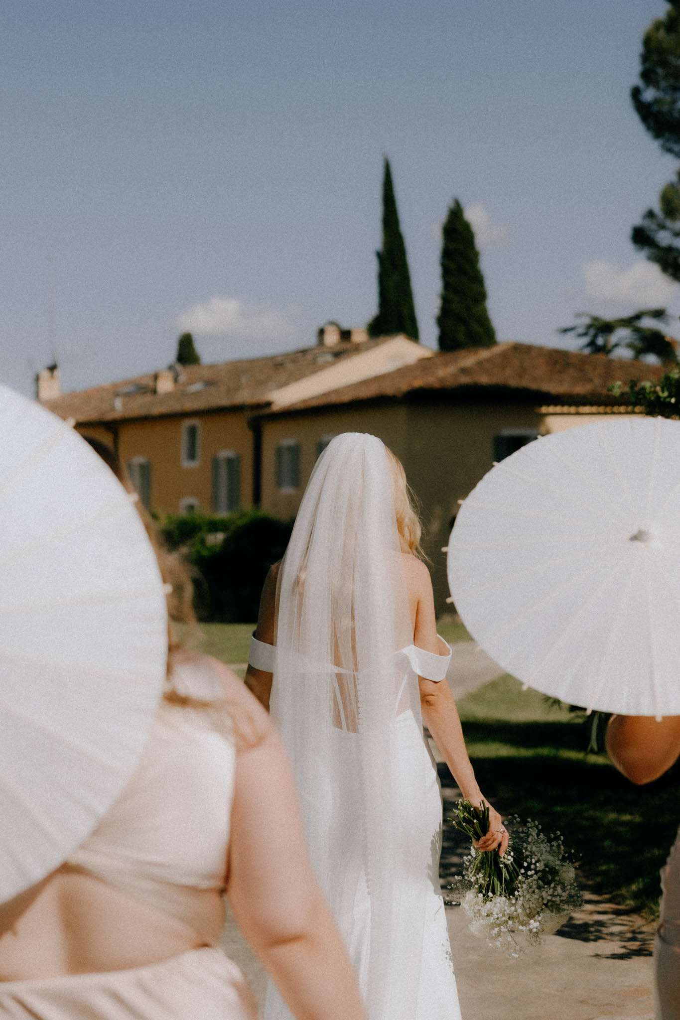 The bride is walking outdoors toward a large ochre-colored villa with a terracotta roof, flanked by at least two attendants holding white paper parasols to shade her from the sun. The bride wears a sleeveless white gown with an off-the-shoulder or strapless neckline and a mid-length plain-edge veil, and carries a loose, organic bouquet of white gypsophila and small delicate blooms. The attendants wear light champagne or blush-toned dresses. The shot is taken from behind at mid-distance, giving a candid, processional feel with the venue building as the backdrop.