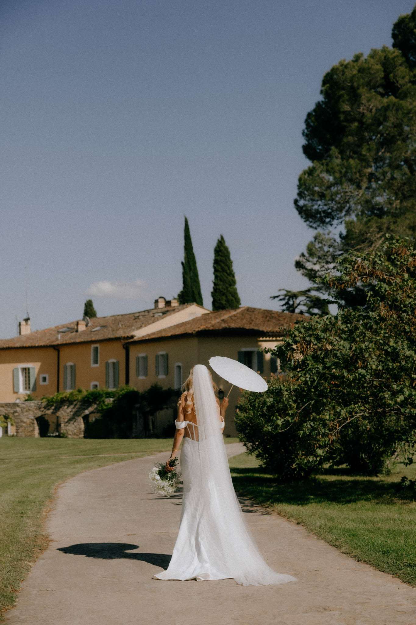 Bride from behind walking gravel path with cathedral veil white parasol and bouquet toward ochre mas