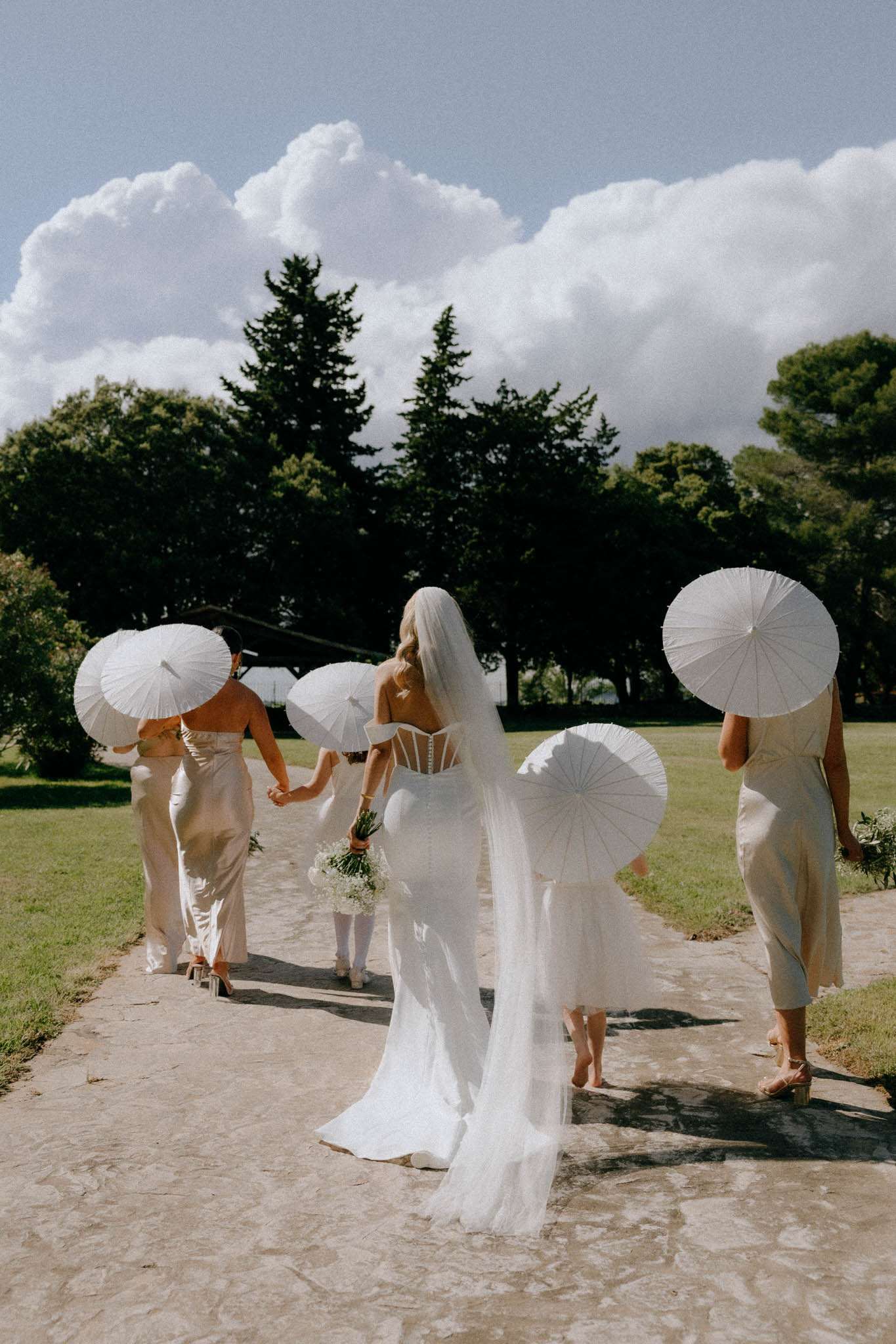 Bride with cathedral veil and babys breath bouquet walking with four bridesmaids in champagne slips holding white parasols