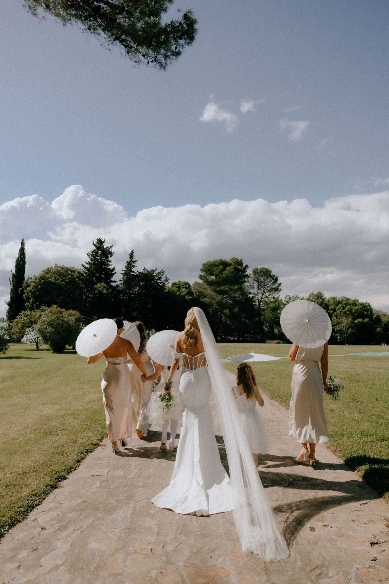 Bride in white off-shoulder gown with veil and three bridesmaids in champagne satin dresses carrying white parasols