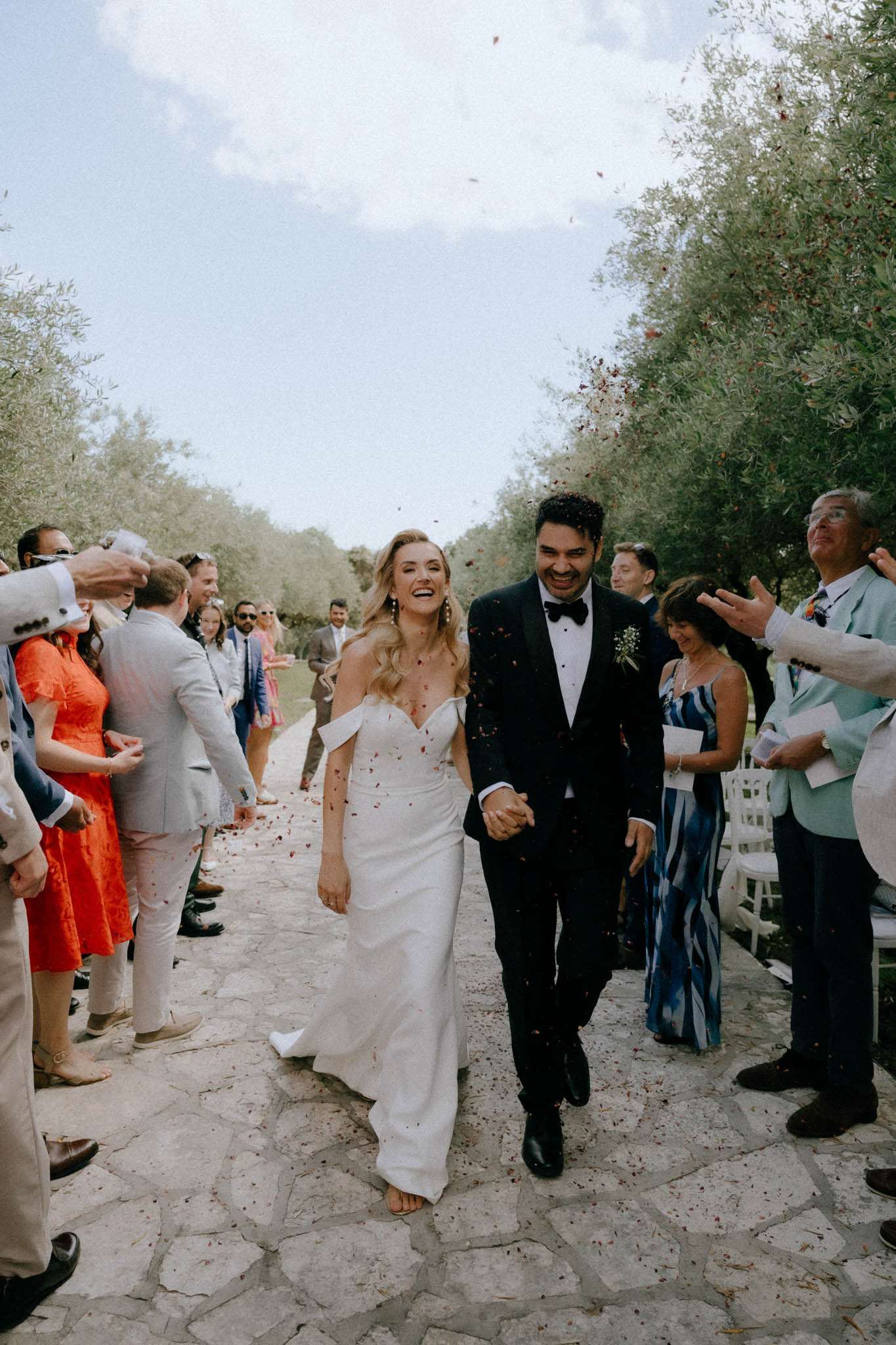 Bride and groom recessional through red petal confetti on olive-tree-lined path with seated guests