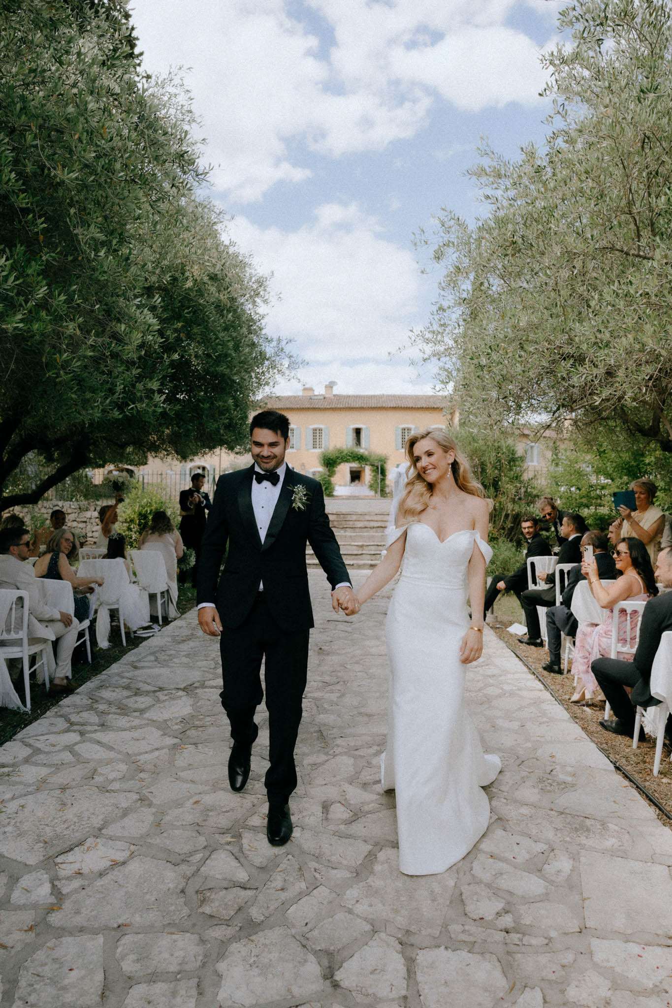 The couple is walking back down the aisle following an outdoor ceremony, holding hands and smiling. The ceremony took place on a flagstone path lined with mature olive trees, with white chairs arranged on either side seating approximately 30–40 guests. The groom wears a black tuxedo with a bow tie and a small boutonniere of greenery and white flowers, while the bride wears an off-the-shoulder fitted white gown with a sweetheart neckline and a short train. A Provençal-style ochre-rendered manor house with shuttered windows and ivy is visible in the background at the top of stone steps, indicating a classic French countryside property. The styling is clean and modern with a black-and-white formal palette, and the seating arrangement and stone pathway create a natural, understated outdoor setting. Wide portrait shot taken at ground level facing the couple.