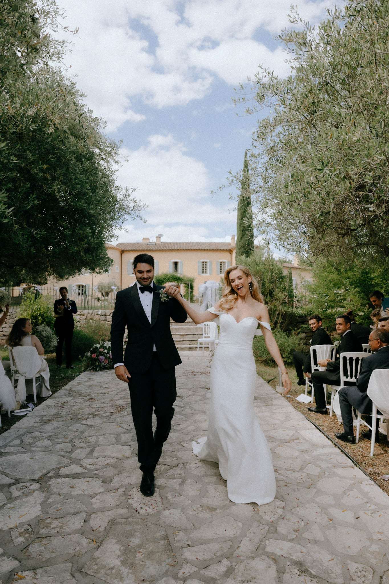 The couple is walking back up the aisle following their outdoor ceremony, with the bride laughing and raising their joined hands in celebration. The setting is a stone-paved pathway at a Provençal property, with a warm ochre-toned stone farmhouse visible in the background. The bride wears an off-the-shoulder white fitted gown with a short train, and the groom wears a black tuxedo with a bow tie and a small floral boutonniere. Guests — approximately 20 visible — are seated on white Napoléon chairs arranged on either side of the aisle, dressed predominantly in dark and white attire. The ceremony setup has a classic, understated style with no visible arch or heavy floral installations. The image is a mid-wide portrait shot taken from ground level along the aisle.