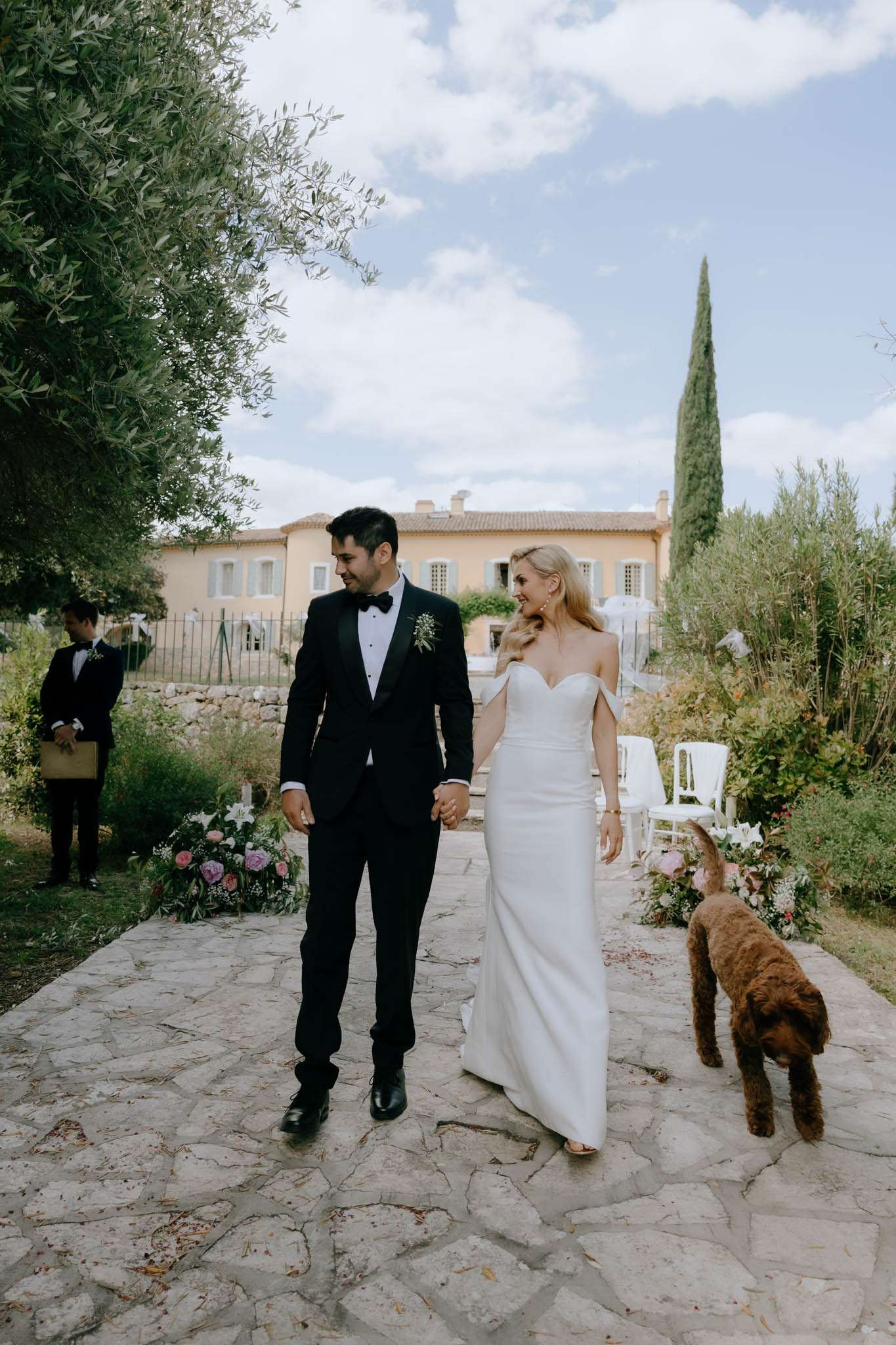 Bride and groom walk hand-in-hand along garden path after ceremony with dog beside them at Provencal mas