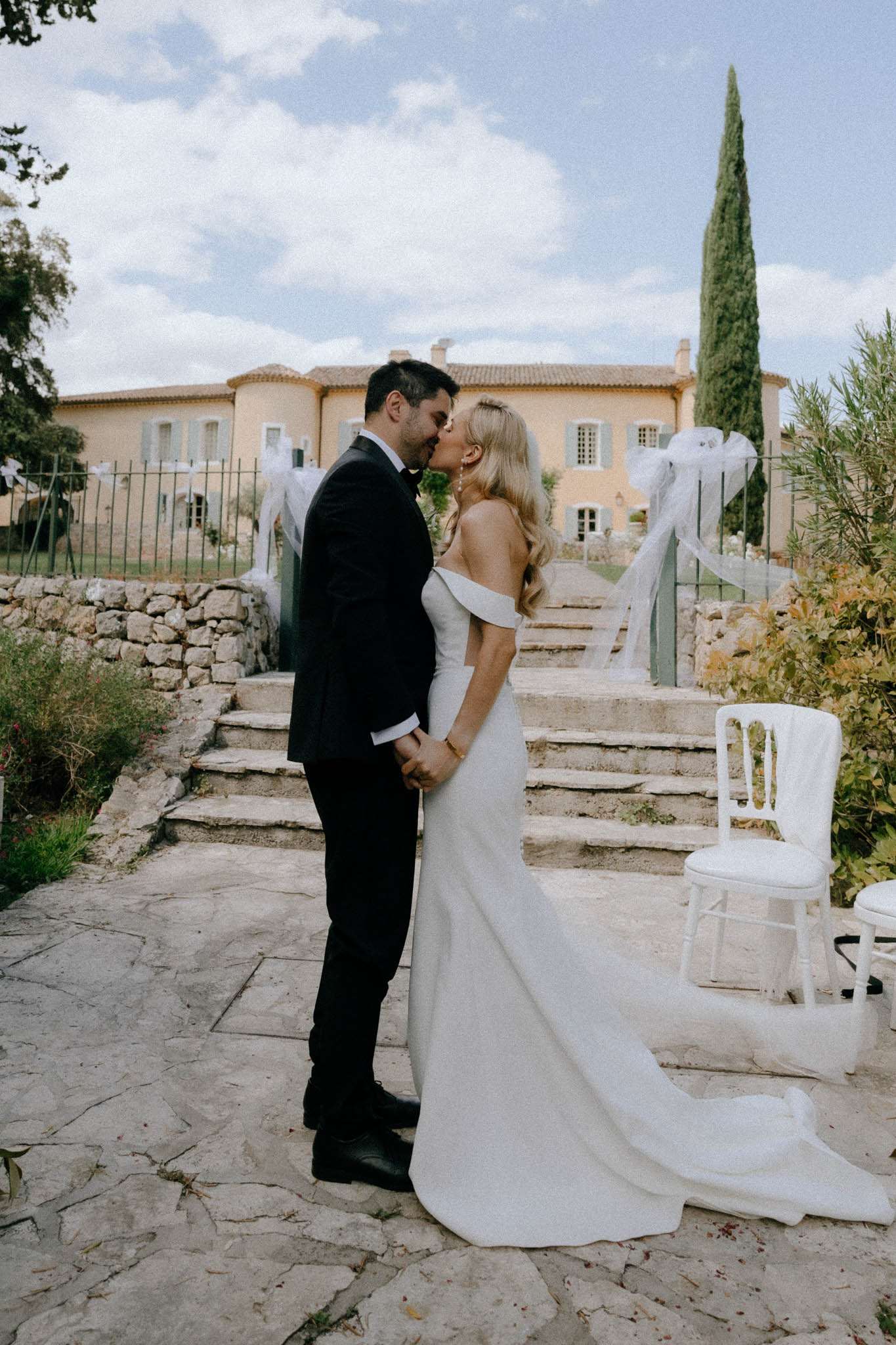 Couple kissing on stone terrace before ochre bastide with sage shutters bride in off-shoulder crepe gown cypress tree behind