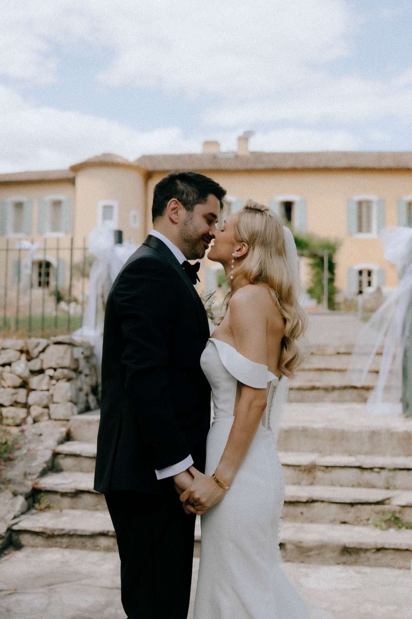 Bride and groom kissing in front of ochre Provencal mas, bride in off-shoulder gown with cathedral veil