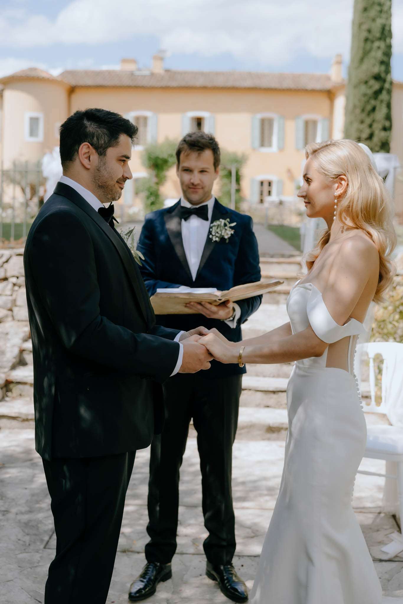 Couple exchanging vows with officiant in navy velvet jacket on Provencal mas terrace with blue shutters