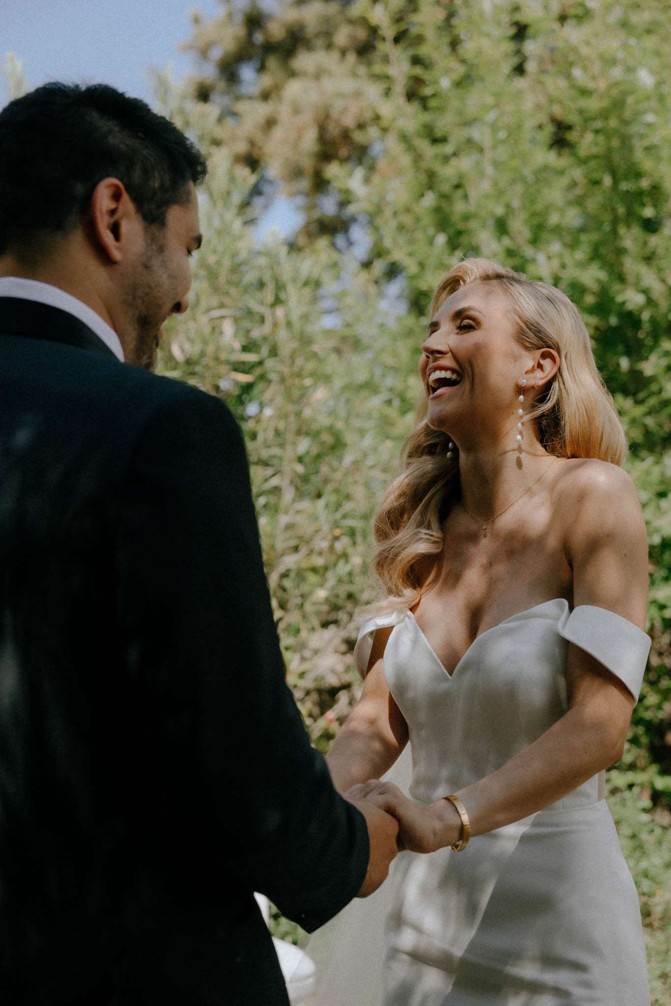 A couple shares a candid, joyful moment outdoors, likely during a first look or outdoor ceremony, with the bride laughing openly while holding the groom's hands. The bride wears an off-the-shoulder ivory satin gown with a structured sweetheart neckline, paired with long pearl drop earrings and a gold bracelet, with her blonde hair styled in loose waves. The groom is seen from behind wearing a dark navy suit. The shot is a medium close-up portrait taken in natural daylight, with a shallow depth of field keeping the couple sharp against a softly blurred green foliage background.