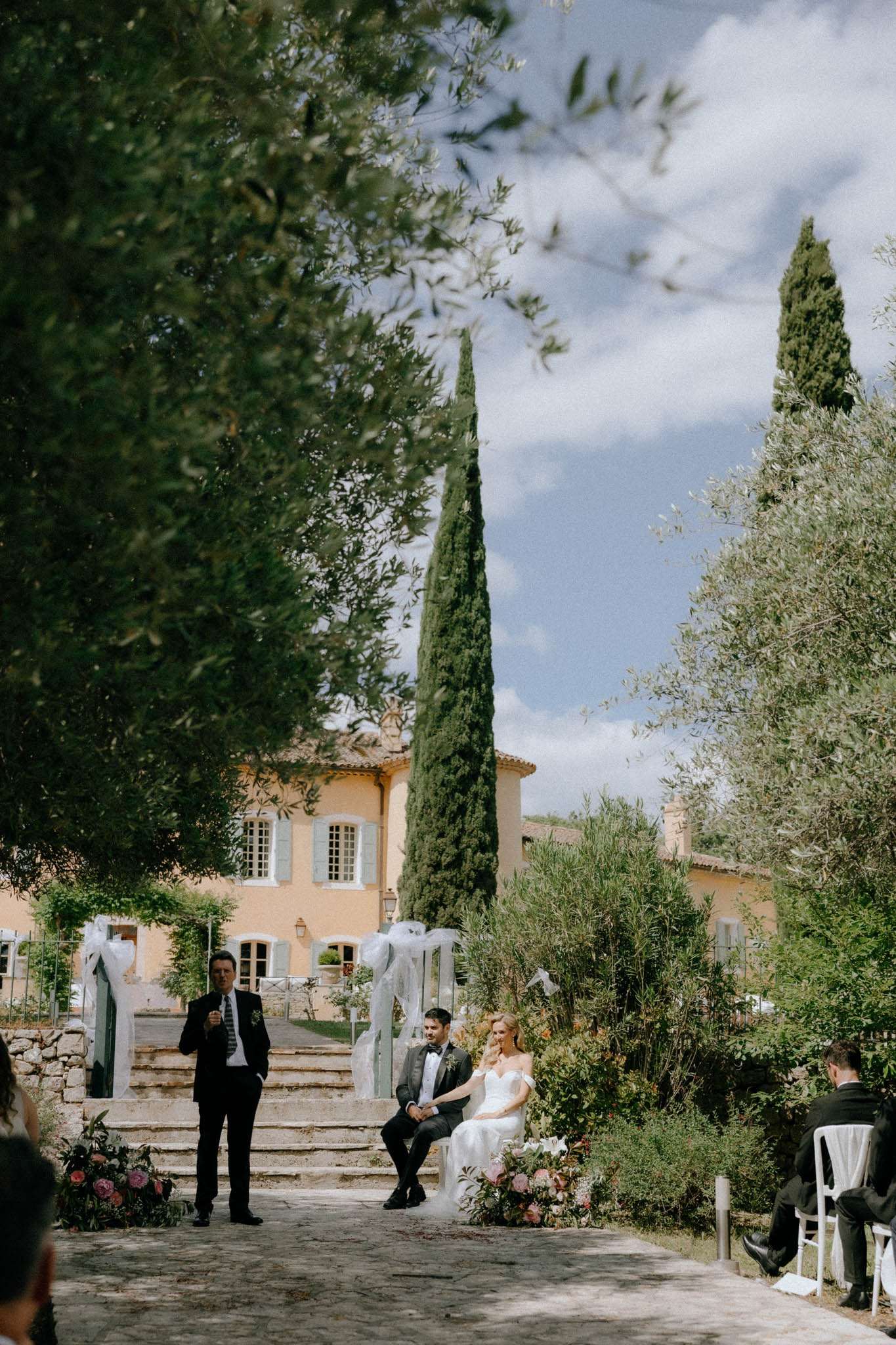 An outdoor wedding ceremony is taking place in the grounds of a Provençal mas or bastide, a ochre-yellow rendered stone property with a terracotta tile roof visible in the background. An officiant in a dark suit stands at a microphone addressing the couple, while the bride in an off-the-shoulder white gown and the groom in a grey suit with black bow tie are seated in front of him on chairs at the altar area. The ceremony backdrop features white fabric-draped pillars flanking stone steps, with floral arrangements of pink and dusty rose blooms at ground level. A small number of guests, partially visible, are seated in white chairs to the right side of the frame. The wide-angle shot is taken from behind tree foliage, framing the scene naturally, with the venue's facade and tall cypress trees prominent in the mid-ground. Potential venue feature image.