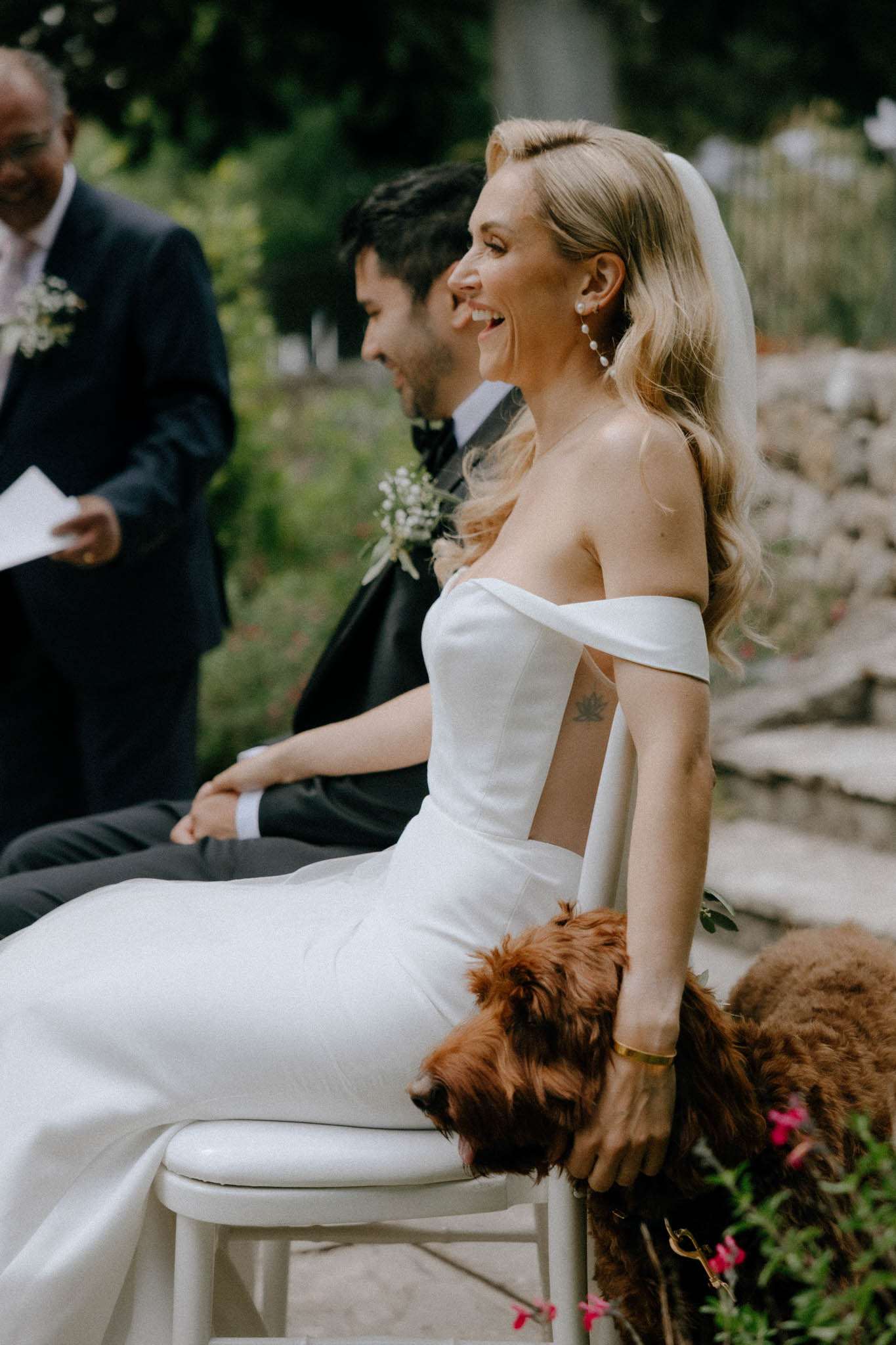 An outdoor ceremony moment capturing the bride and groom seated side by side, laughing and smiling during what appears to be a reading or speech by an officiant or guest visible on the left holding papers. The bride wears a fitted off-the-shoulder white gown with a simple veil and long wavy blonde hair, accessorized with pearl drop earrings and a gold bangle bracelet. The groom is in a black tuxedo with a black bow tie and a small white floral boutonniere. A curly auburn-coated dog is pressed up against the bride's legs beside her white chair. The shot is a close-up portrait taken from a side angle, with a shallow depth of field that blurs the background greenery and stone wall.