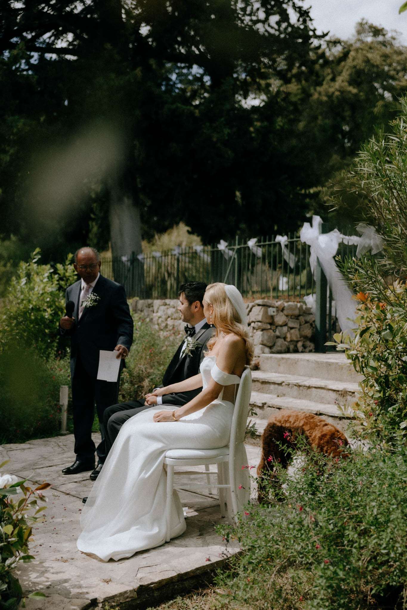 Bride in white gown with pillbox headpiece and groom in tuxedo seated at garden ceremony with officiant