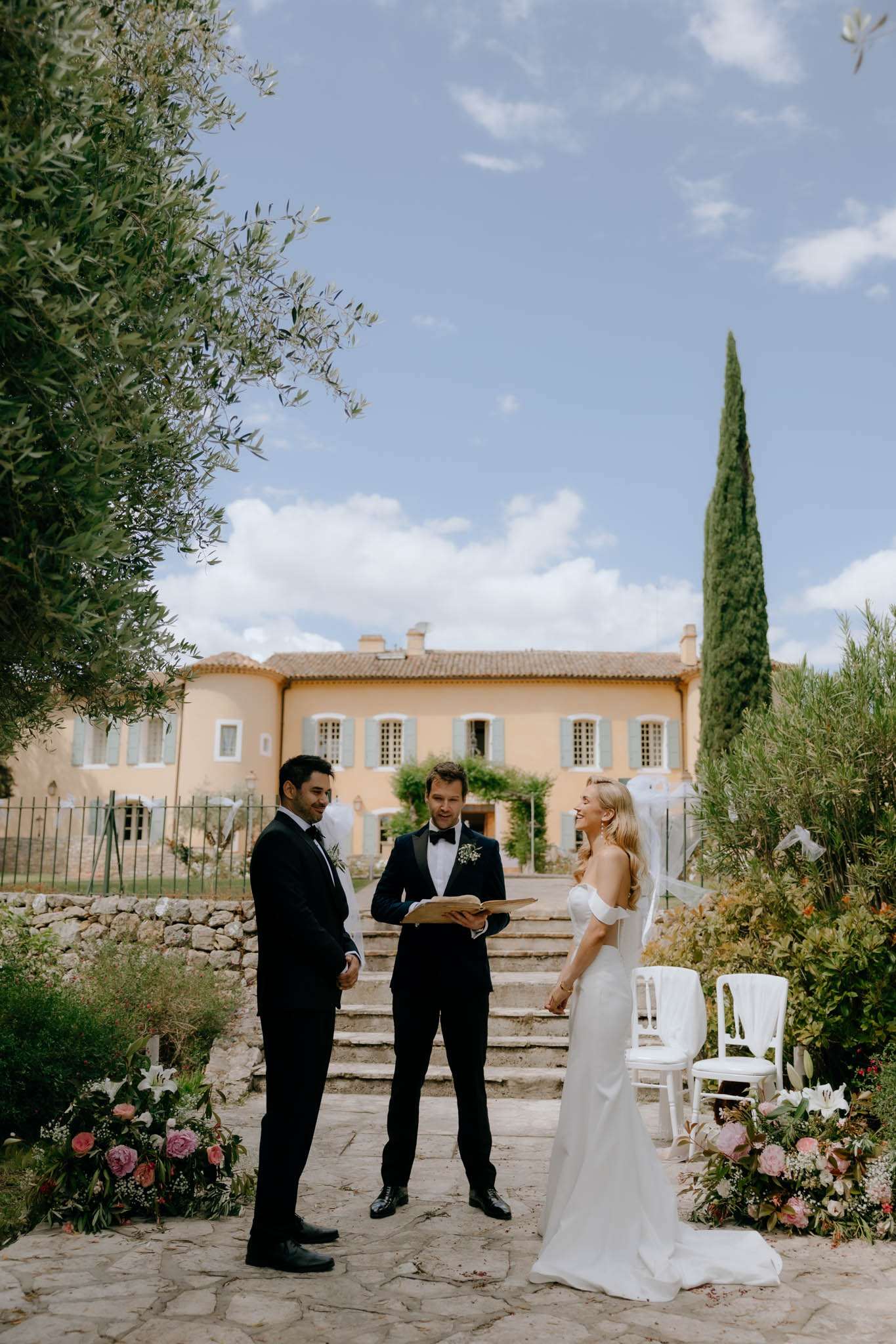 Outdoor ceremony on stone terrace of Provencal manor with bride, groom, officiant, and floral arrangements