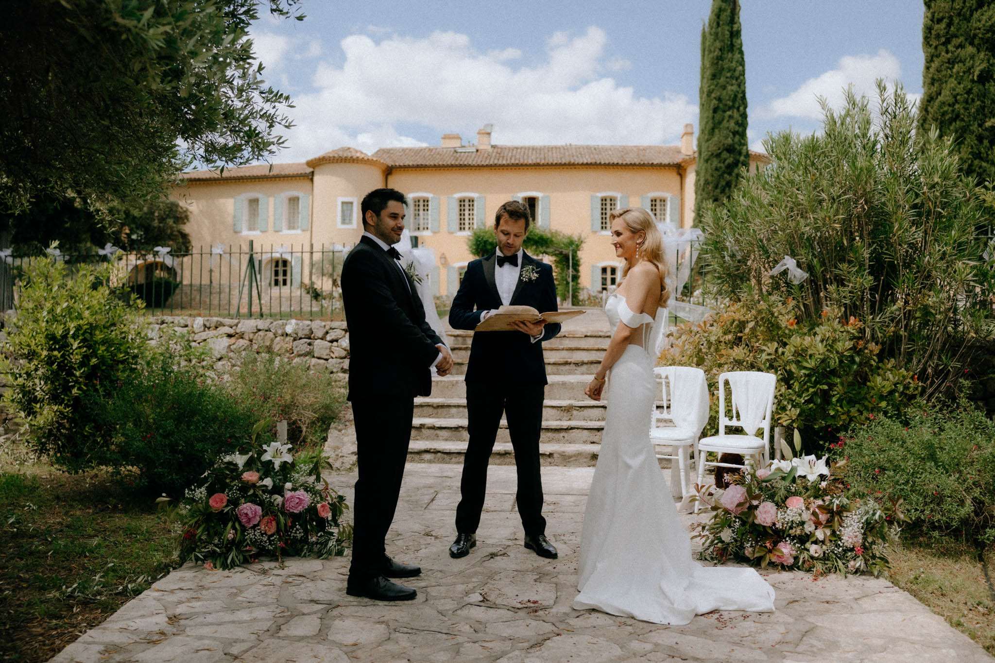 An outdoor wedding ceremony taking place on a stone-paved terrace in front of a Provençal-style manor house with ochre-yellow rendered walls and pale blue shutters. The couple — a groom in a black tuxedo with a white floral boutonnière and a bride in an off-the-shoulder fitted white gown with a cathedral-length veil — face each other while an officiant in a navy tuxedo reads from a leather-bound book between them. The ceremony space is flanked by low floral ground arrangements featuring pink peonies, white lilies, and lush greenery, with white chiavari chairs visible to the right. Stone steps lead up toward the manor behind the officiant, with tall cypress trees framing the venue. The overall styling is classic with a romantic garden feel, captured in a wide environmental portrait shot.