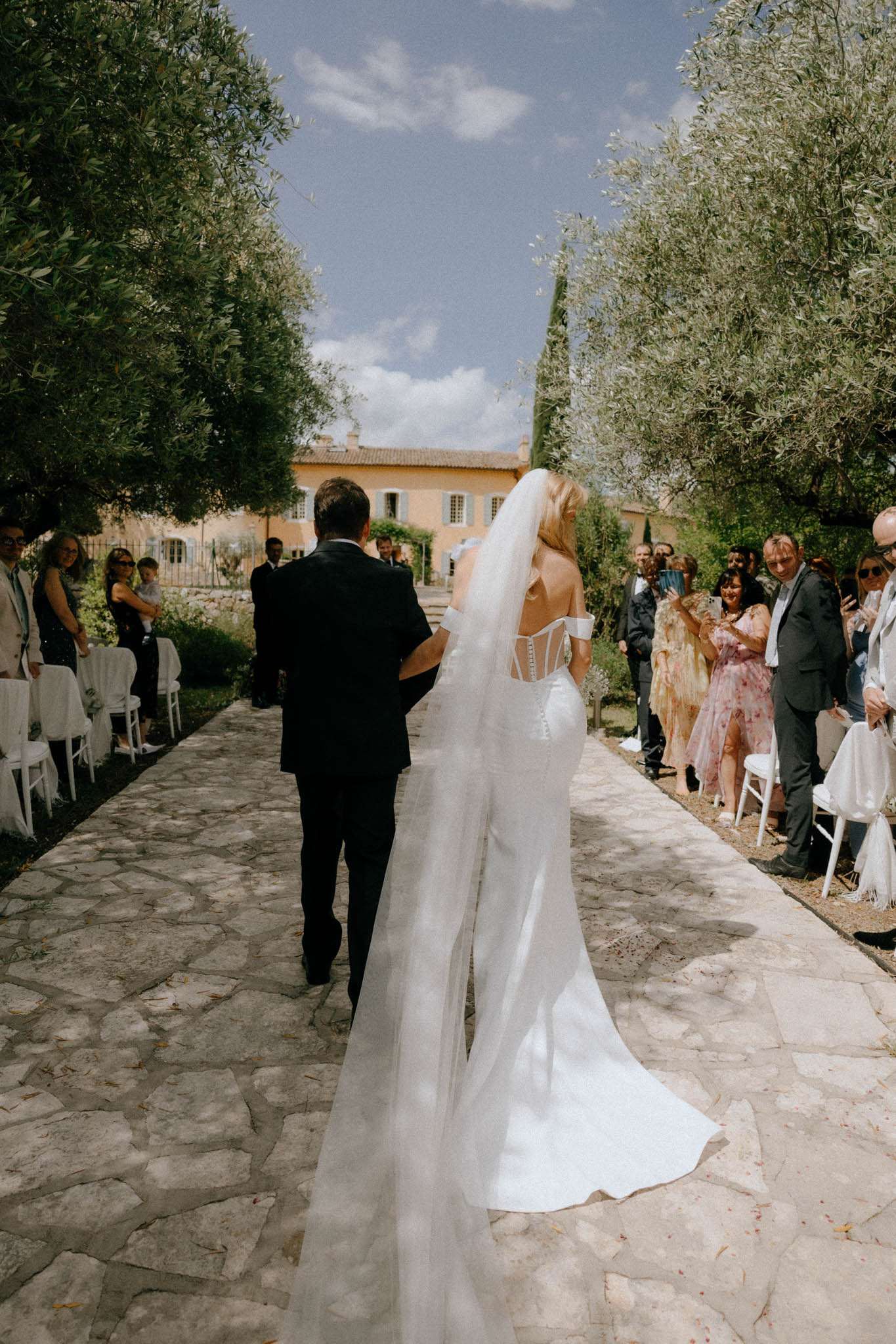 Bride walked down stone-paved outdoor aisle from behind showing cathedral veil and corset gown at Provencal venue