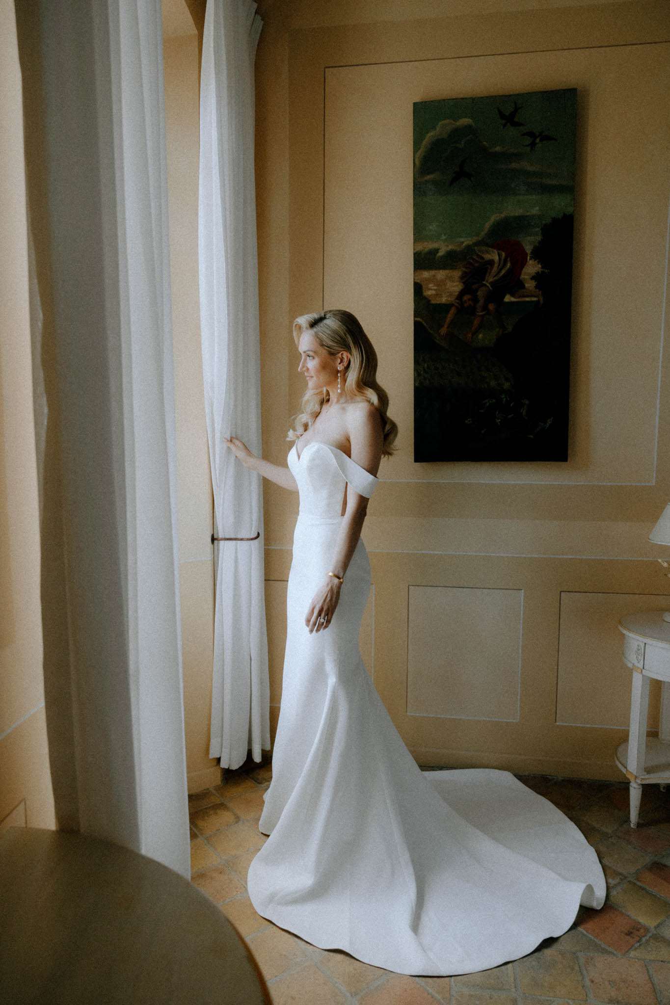 A bridal portrait taken indoors in what appears to be a room within a French château or villa, featuring warm ochre-toned walls, terracotta tile flooring, and a large classical oil painting on the wall. The bride stands in profile near a tall window with white linen curtains, gazing outward and holding the curtain panel lightly with one hand. She wears a white off-the-shoulder fitted gown with a structured sweetheart neckline and a floor-length train pooling behind her. Her accessories include drop pearl earrings, a gold bracelet, and a ring, and her blonde hair is styled in loose waves swept to one side. The shot is a full-length portrait with warm, natural window light illuminating her from the side.