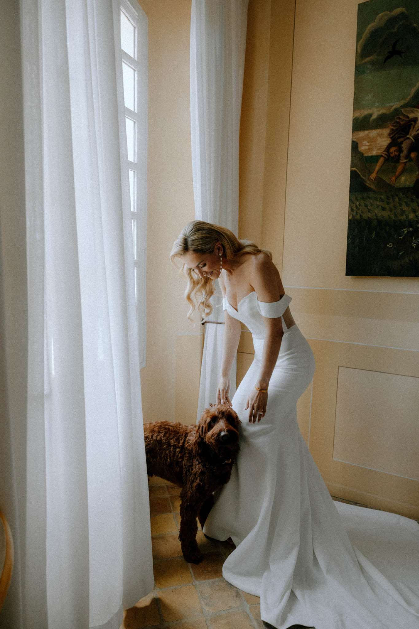 Bride in off-the-shoulder white gown with pearl earrings bends to pet a brown dog in a chateau interior