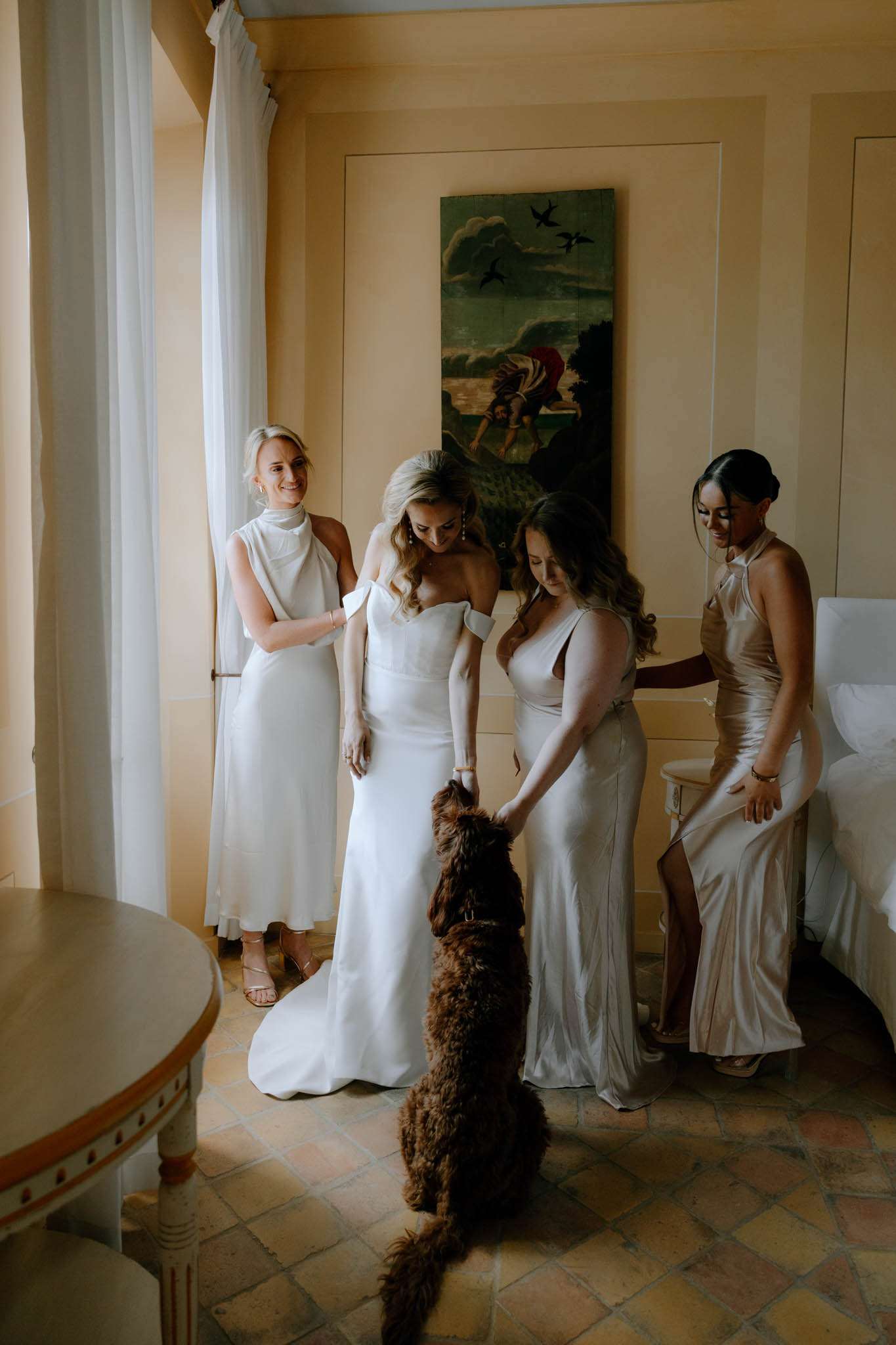 Bride in white off-shoulder gown and bridesmaids in champagne satin dresses interacting with curly-haired brown dog indoors