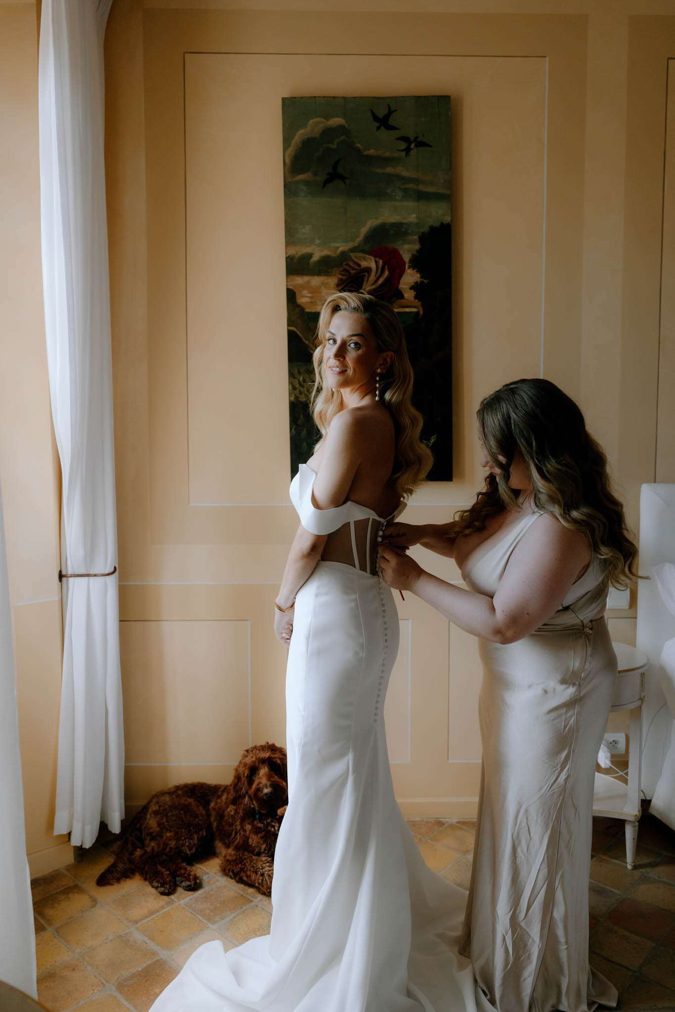 Bridesmaid fastening the back of the bride's white corset gown in a peach-toned room with a dog resting nearby