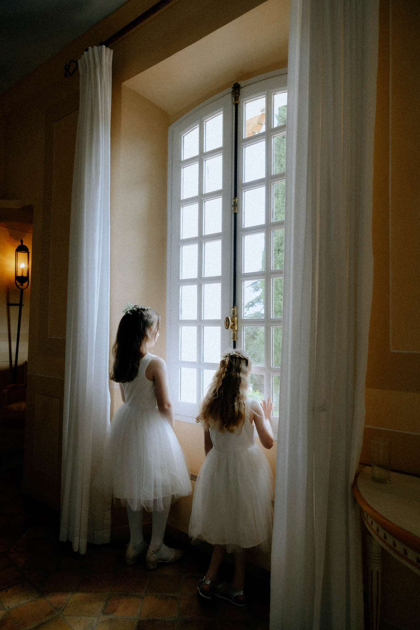Two flower girls in white tulle dresses with floral crowns looking out through an arched chateau window