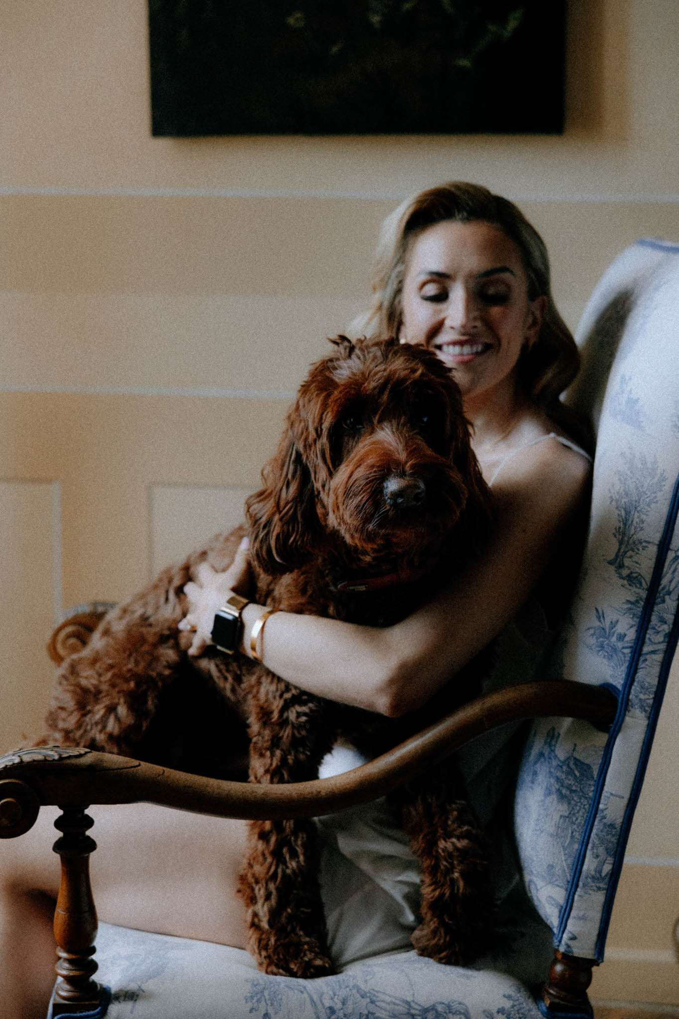 Bride in strapless gown laughing with curly-coated dog in toile armchair in chateau interior