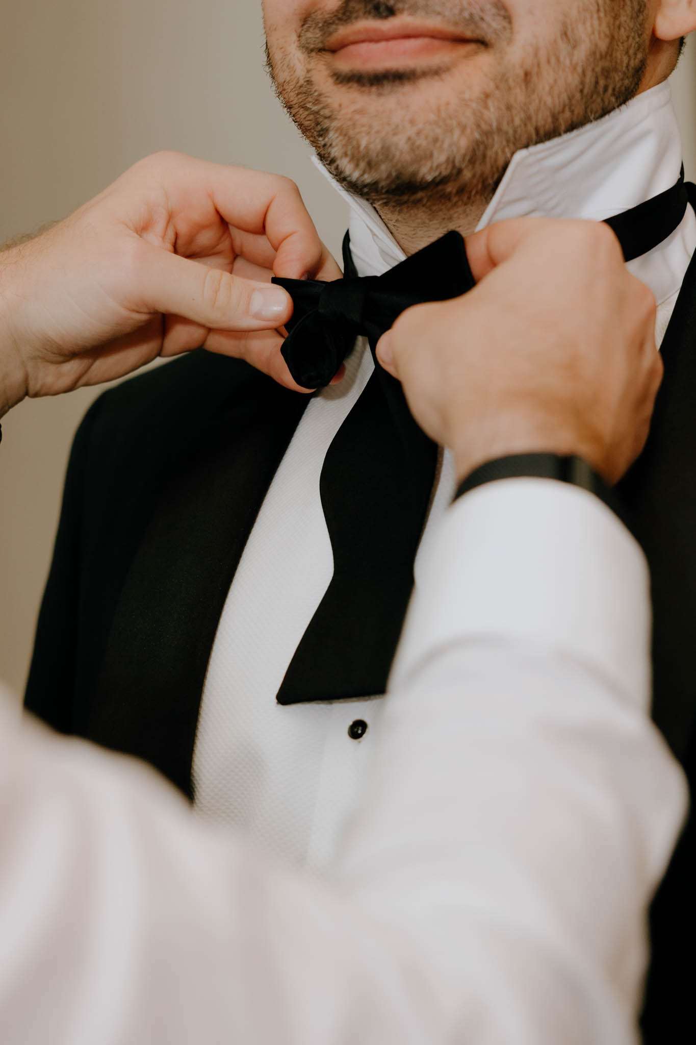 Hands adjusting black velvet bow tie on groom's tuxedo with satin lapels and white pique dress shirt