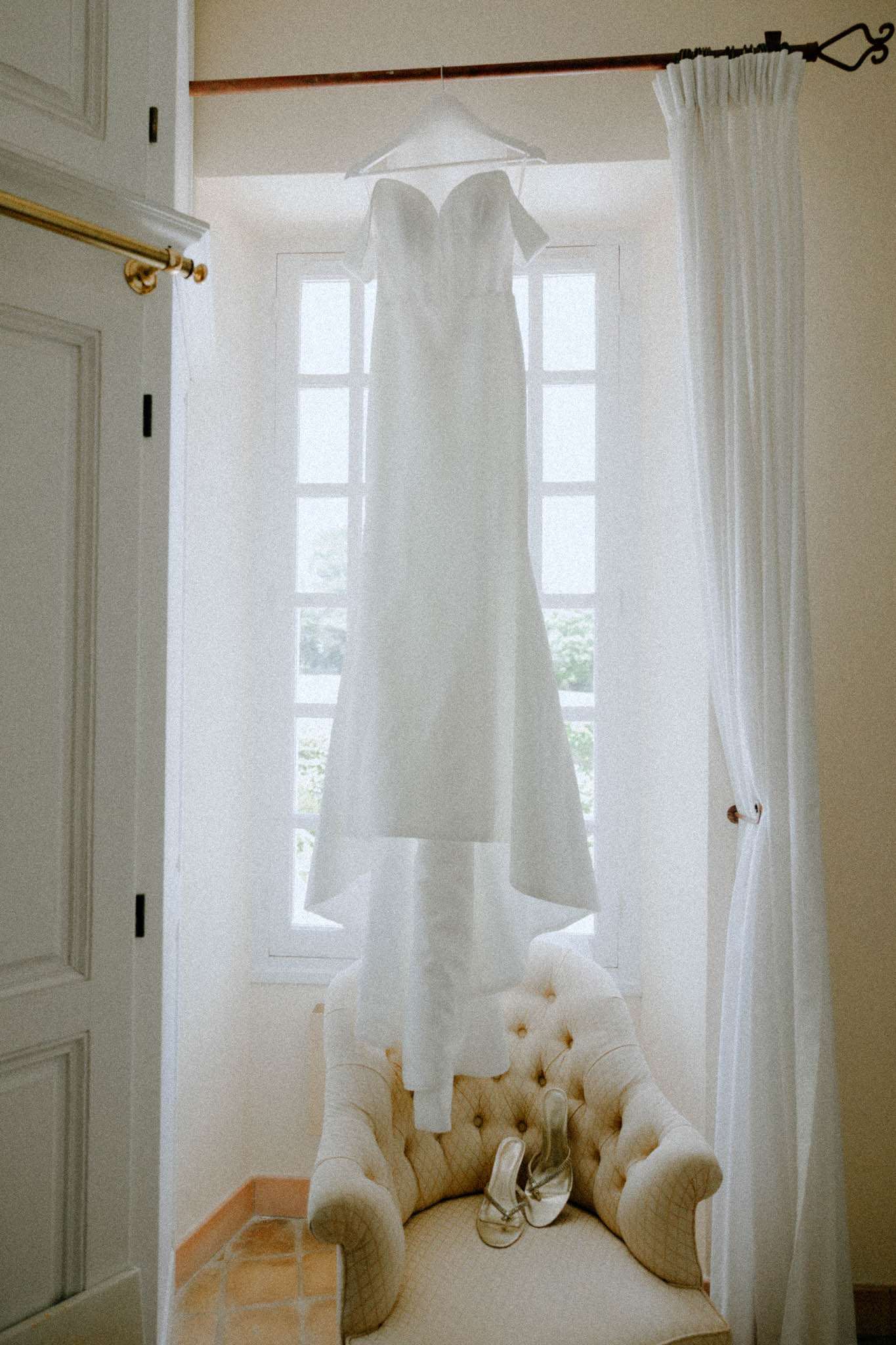 White strapless wedding dress hanging by a window with silver heeled sandals on a cream armchair in a French chateau room