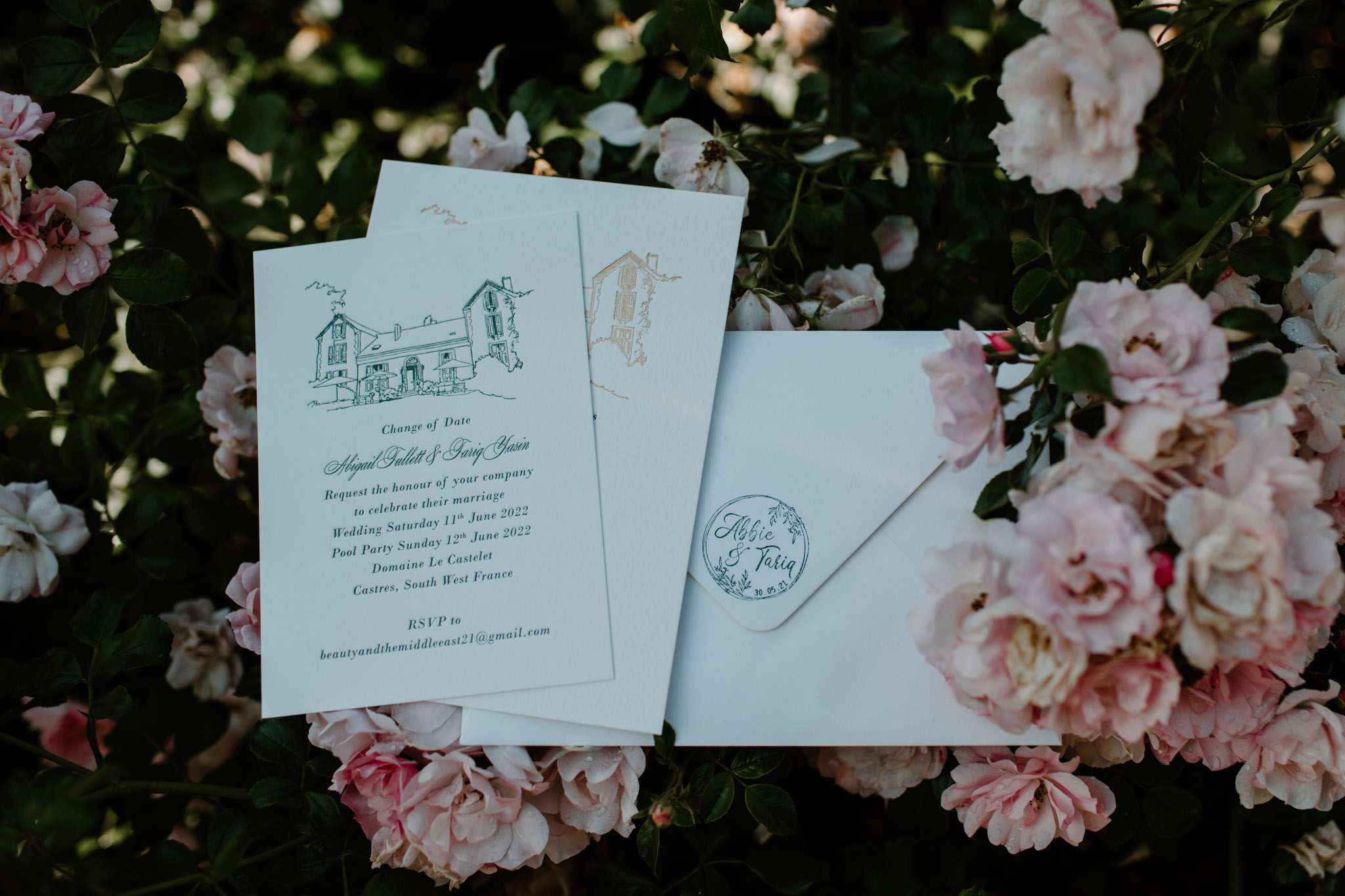 A close-up flat lay detail shot of a wedding stationery suite placed among blooming pale pink roses. The suite includes a white invitation card featuring a hand-drawn ink illustration of the venue building at the top, printed in dark green, with script and serif typography announcing a change of date for Abigail Tullett and Tariq Yasin, with the wedding on Saturday 11th June 2022 and a Pool Party on Sunday 12th June 2022 at Domaine Le Castelet, Castres, South West France. Behind the invitation is a second card showing a gold-toned version of the same venue illustration. A white envelope sits alongside, sealed with a circular wax or paper seal embossed with the names 'Abbie & Tariq' and a floral wreath motif. The overall stationery palette is white and soft green with gold accents, styled in a classic illustrative design.