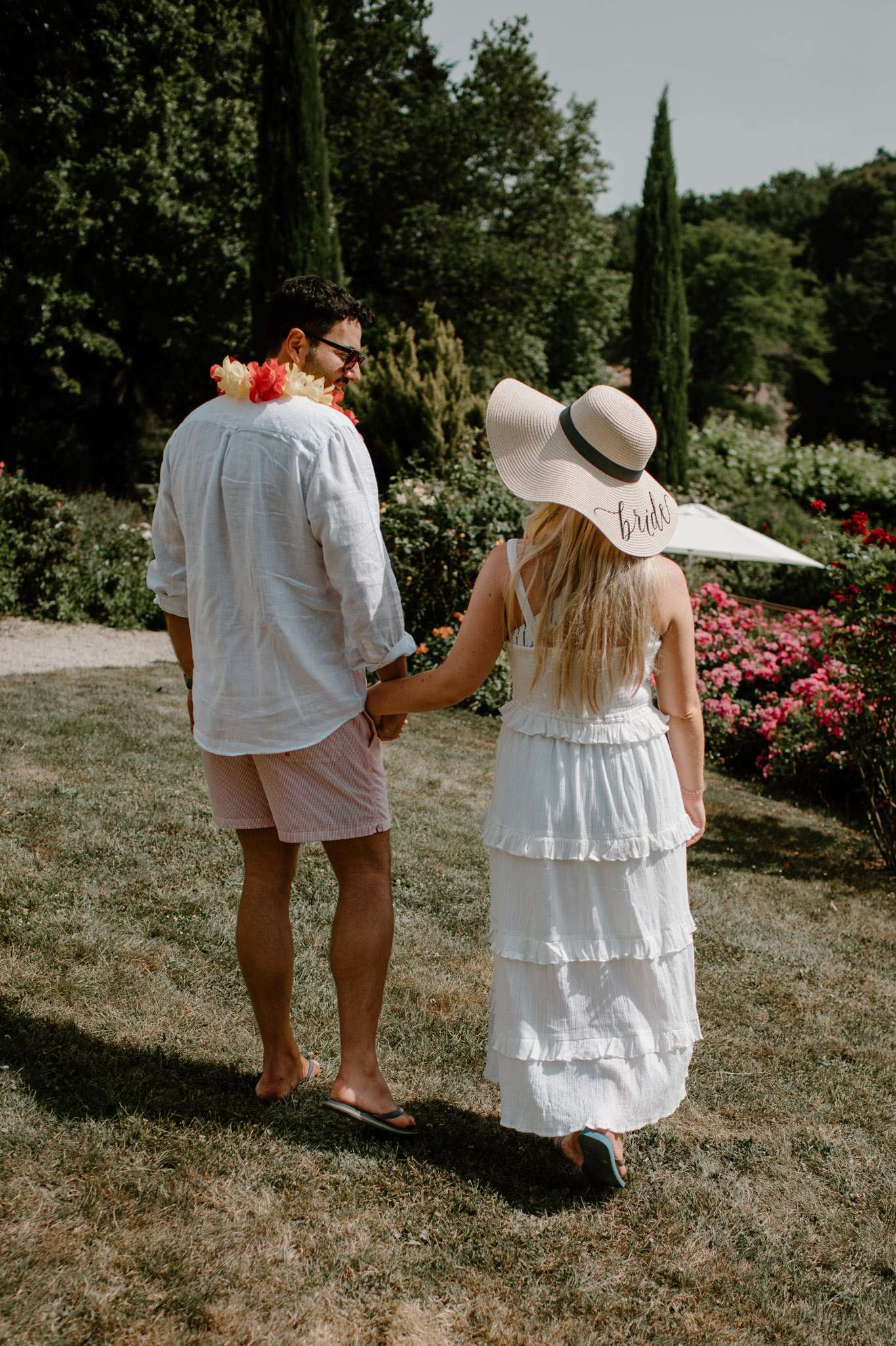 A couple walks hand-in-hand through a garden during what appears to be a pre-wedding or wedding weekend casual event. Both are photographed from behind in a medium full-body shot. The woman wears a white tiered ruffle maxi dress, light blue sandals, and a wide-brim white sun hat with a black band embroidered with the word 'bride' in script. The man wears a white linen shirt, pink shorts, flip flops, and a red and yellow tropical flower lei around his neck. The outdoor garden setting features vibrant pink rose bushes, cypress trees, and a gravel path visible to the left, suggesting a French estate or château property.