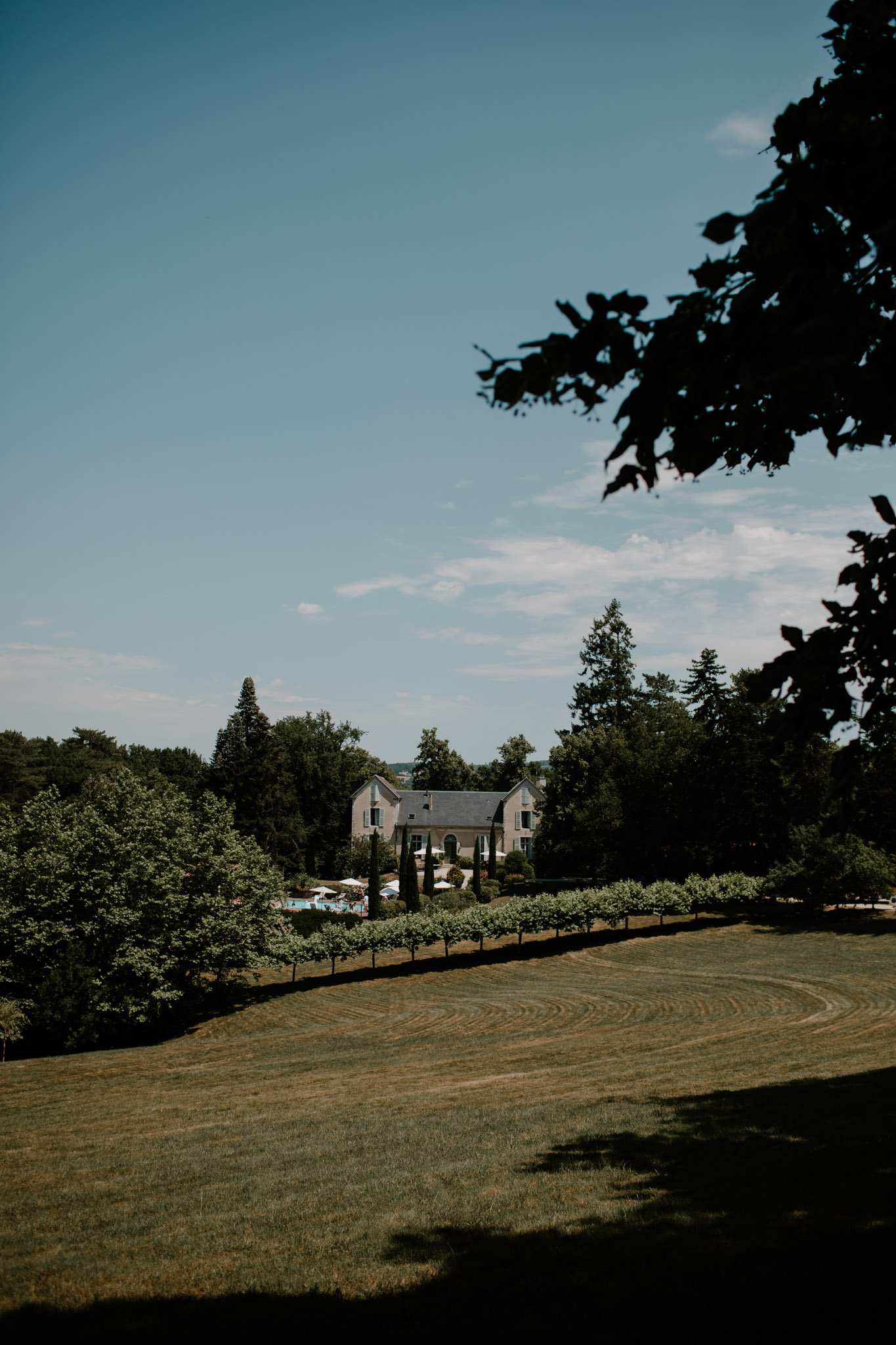 Wide landscape view of a French manor house surrounded by mature trees with white parasols on the terrace