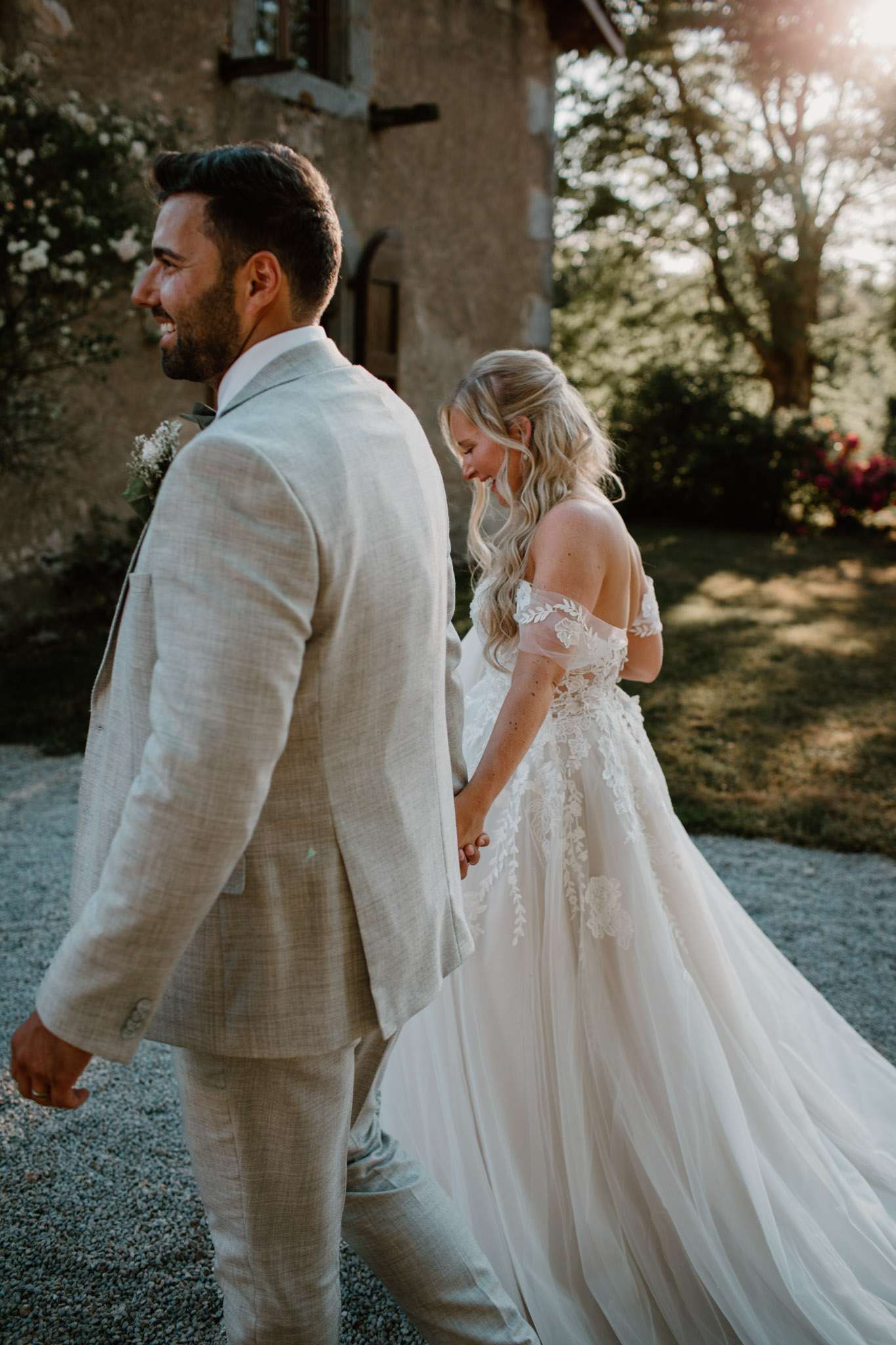 A couple portrait taken outdoors on a gravel path beside a stone building, shot during golden hour with warm backlit sun flare. The groom walks slightly ahead, smiling, wearing a light beige linen suit with a white and greenery boutonniere. The bride follows, looking downward with a soft smile, wearing an off-the-shoulder ivory ball gown with floral lace appliqué detailing on the bodice and a full tulle skirt; her blonde wavy hair is worn half-up. The two are holding hands mid-stride. The overall styling aesthetic is romantic boho with a relaxed, natural feel. Medium-distance portrait shot with a shallow depth of field.