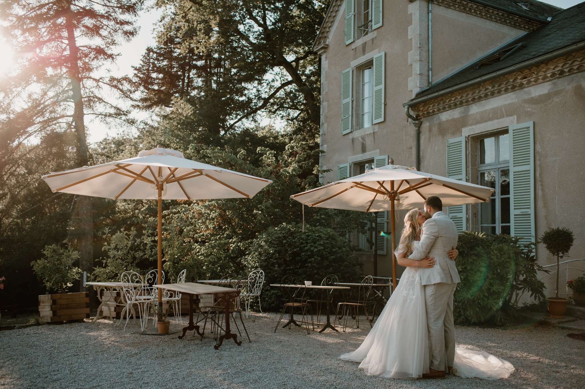 A couple shares a kiss during an outdoor couple portrait on the gravel terrace of a French château, with warm golden-hour sunlight casting a soft lens flare across the scene. The bride wears a white ballgown with lace detailing and a long train, while the groom is dressed in a light grey suit. The terrace is set with wooden bistro tables, white wrought-iron chairs, darker metal café chairs, and two large white market umbrellas with wooden frames, suggesting a cocktail hour setup. The château façade features sage green shutters and ornate stone detailing, visible to the right of the frame in this wide environmental portrait. Potential venue feature image.
