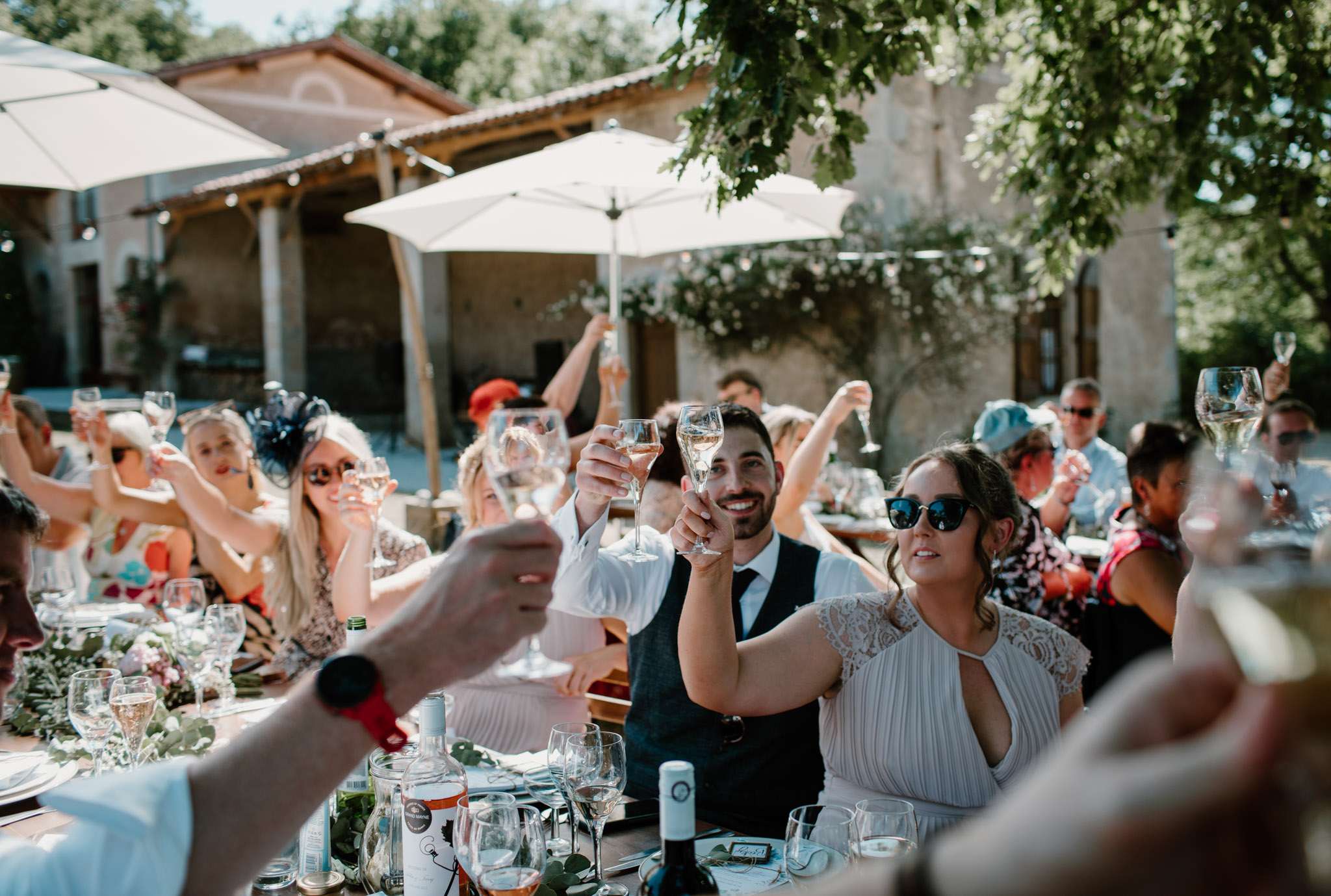 25 guests raising glasses during toast at communal table under umbrellas at rustic stone domaine
