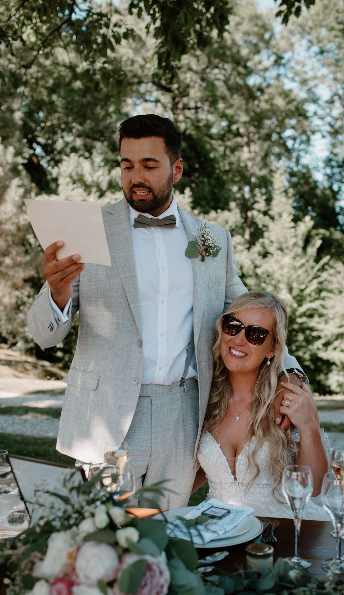 Groom reading speech from card at outdoor reception table while bride sits beside him smiling