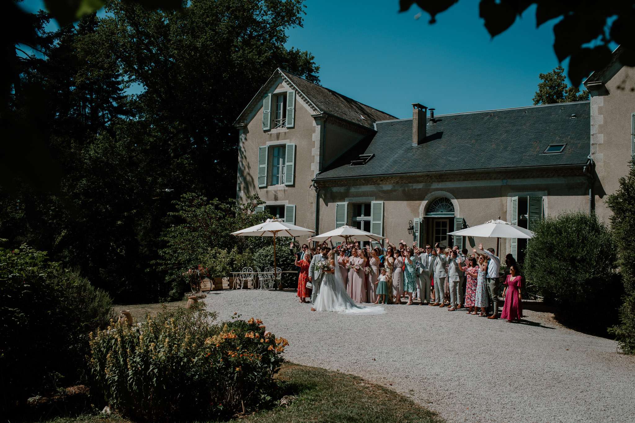 Large wedding group photo with arms raised on gravel courtyard in front of French manor house