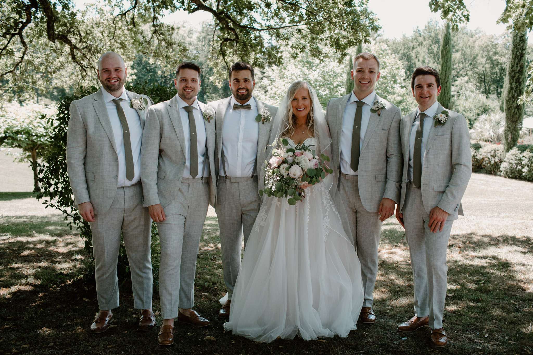 Bride in lace ballgown with peony bouquet posed with groom and three groomsmen in grey suits outdoors