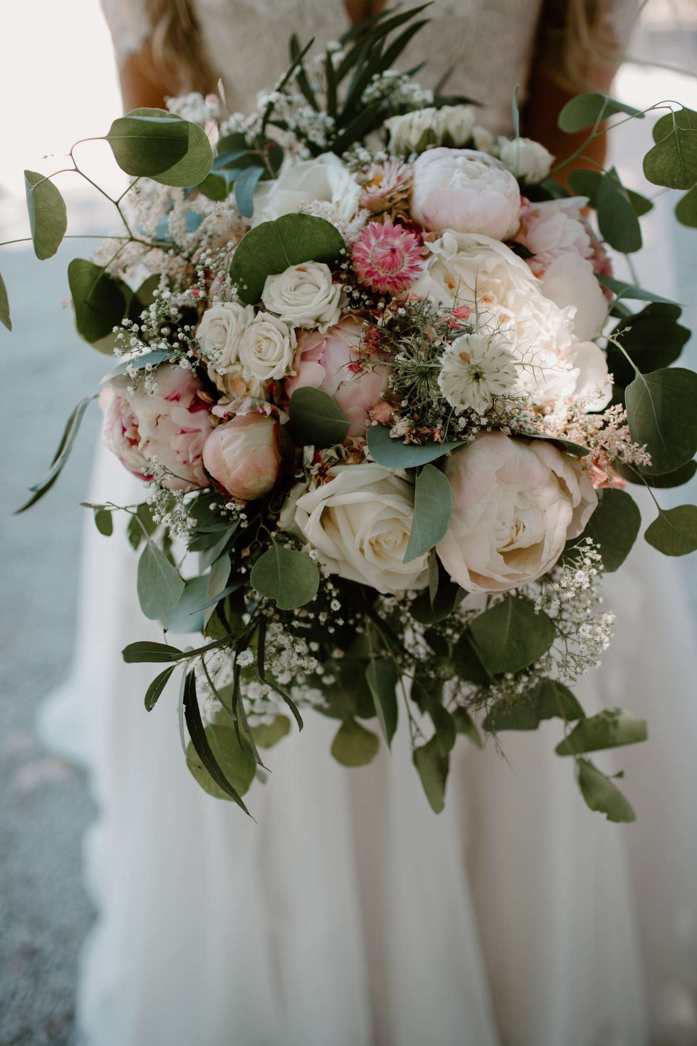 Close-up of bridal bouquet with blush peonies, cream roses, strawflowers, and trailing eucalyptus held by bride in white l...