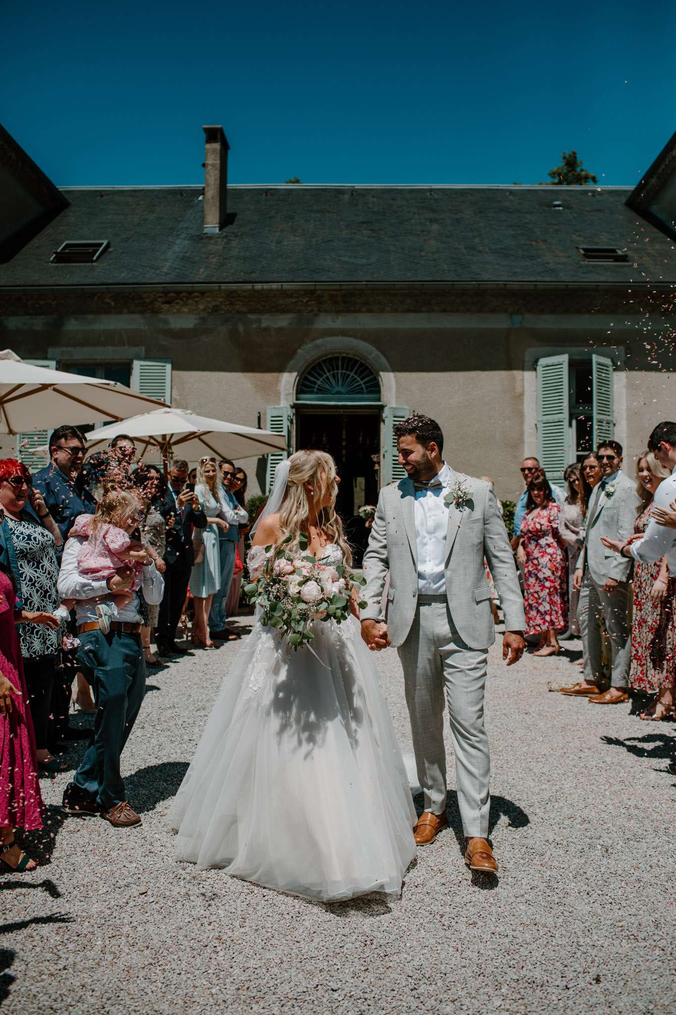 Bride and groom walking through pink petal confetti exit before chateau with blue shutters