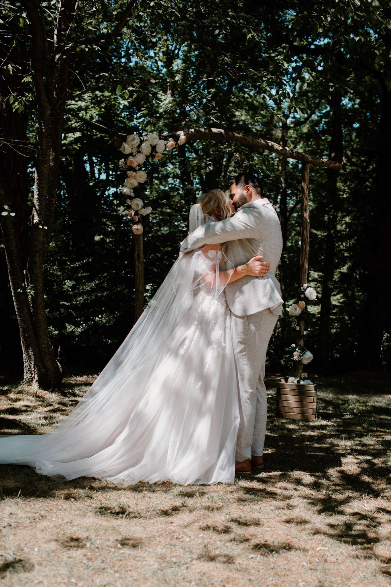 Bride and groom share first kiss under a rustic branch arch decorated with cream and blush roses