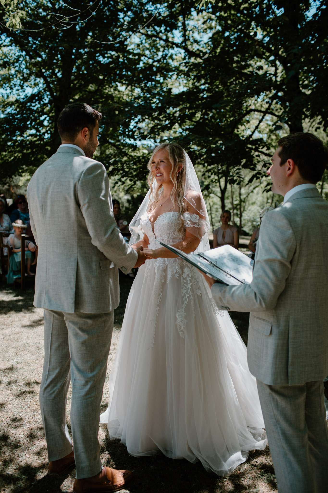An outdoor wedding ceremony taking place beneath large shade trees, with the couple exchanging vows while facing each other and holding hands. The bride wears an ivory ballgown with a lace appliqué bodice featuring off-shoulder floral lace detailing and a full tulle skirt, paired with a cathedral-length veil; she has long blonde wavy hair. The groom wears a light grey suit with tan leather shoes. An officiant stands to the right holding a blue binder, also dressed in a light grey suit. Seated guests are visible in the background, with several wearing teal and blue outfits. The overall styling is relaxed and romantic with a garden-boho aesthetic. Medium-distance portrait shot capturing all three standing figures from head to toe.