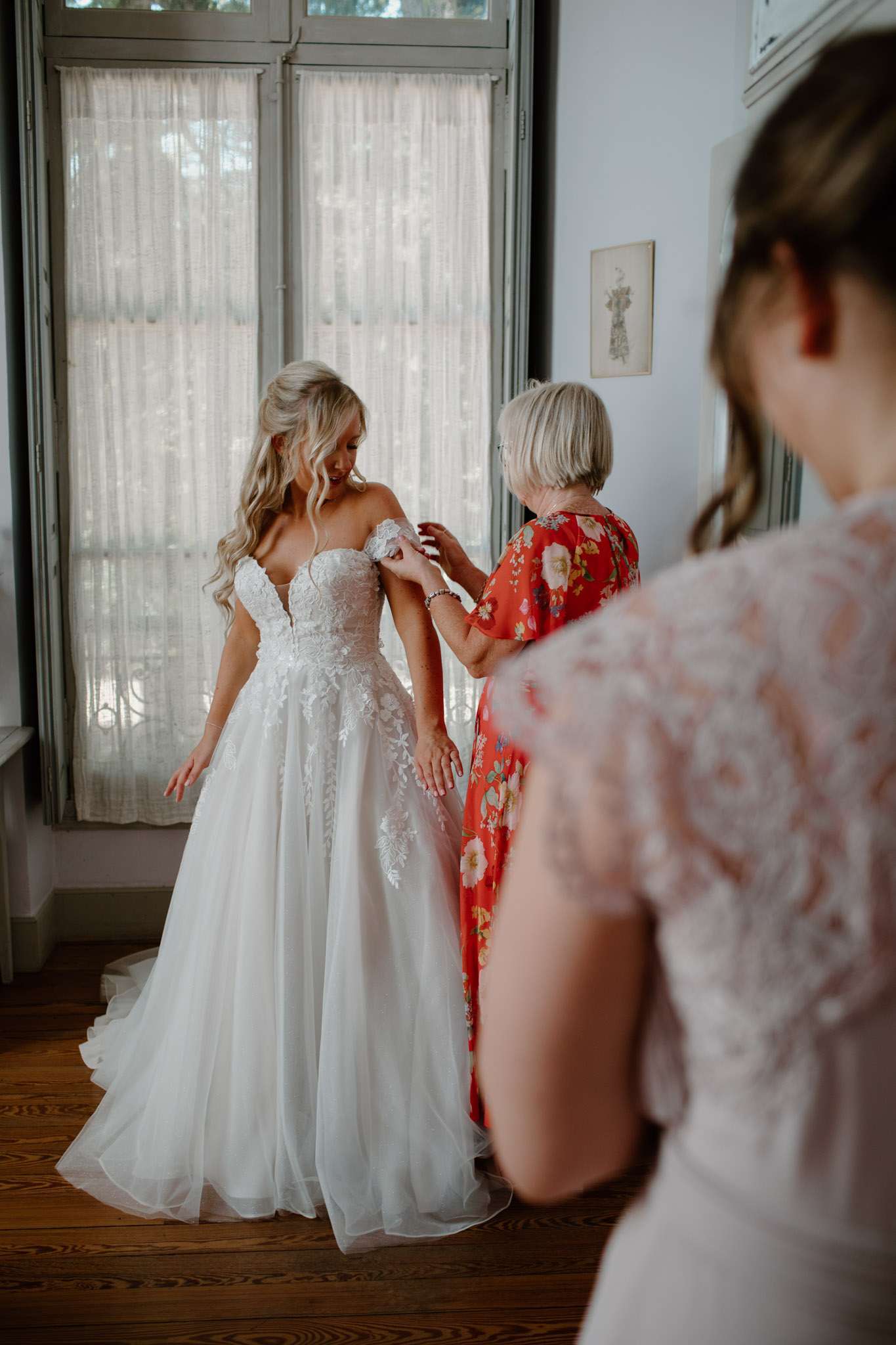 Mother adjusting bride's off-shoulder lace ballgown strap in chateau bridal suite with tall windows