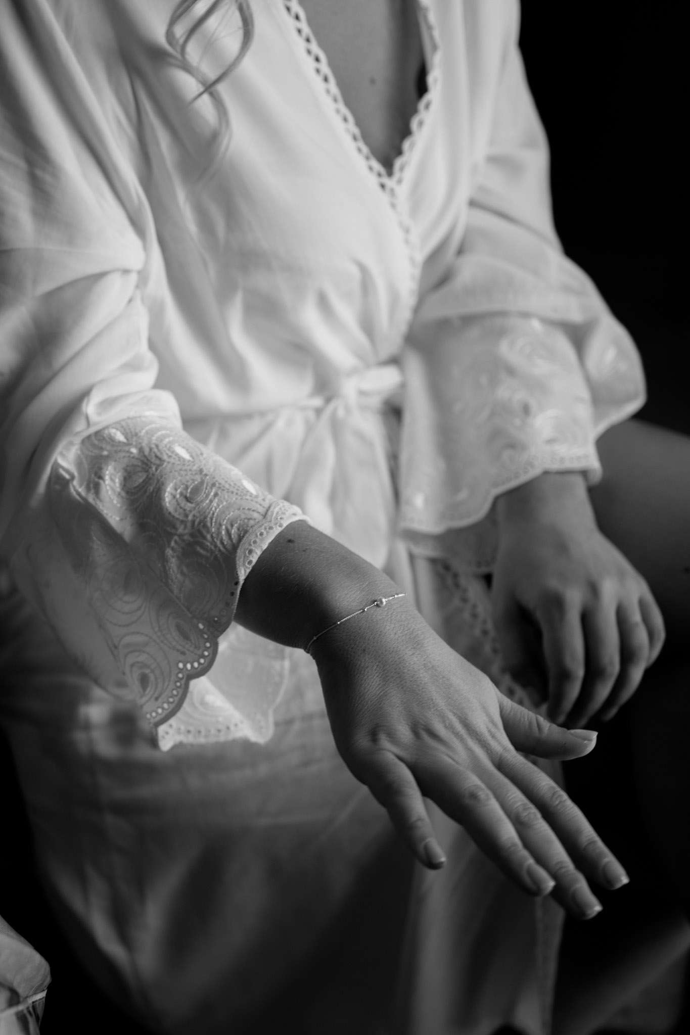 A black-and-white close-up detail shot taken during the getting-ready portion of a wedding day, showing a bride seated and wearing a white robe with broderie anglaise lace cuffs and a tied waist sash. The focus is drawn to her wrist, where a delicate thin chain bracelet with a small charm is displayed. Both hands are visible in the frame, with the left hand extended toward the camera. The high-contrast lighting creates deep shadows against a dark background, emphasizing the texture of the lace sleeve and the fine detail of the bracelet.